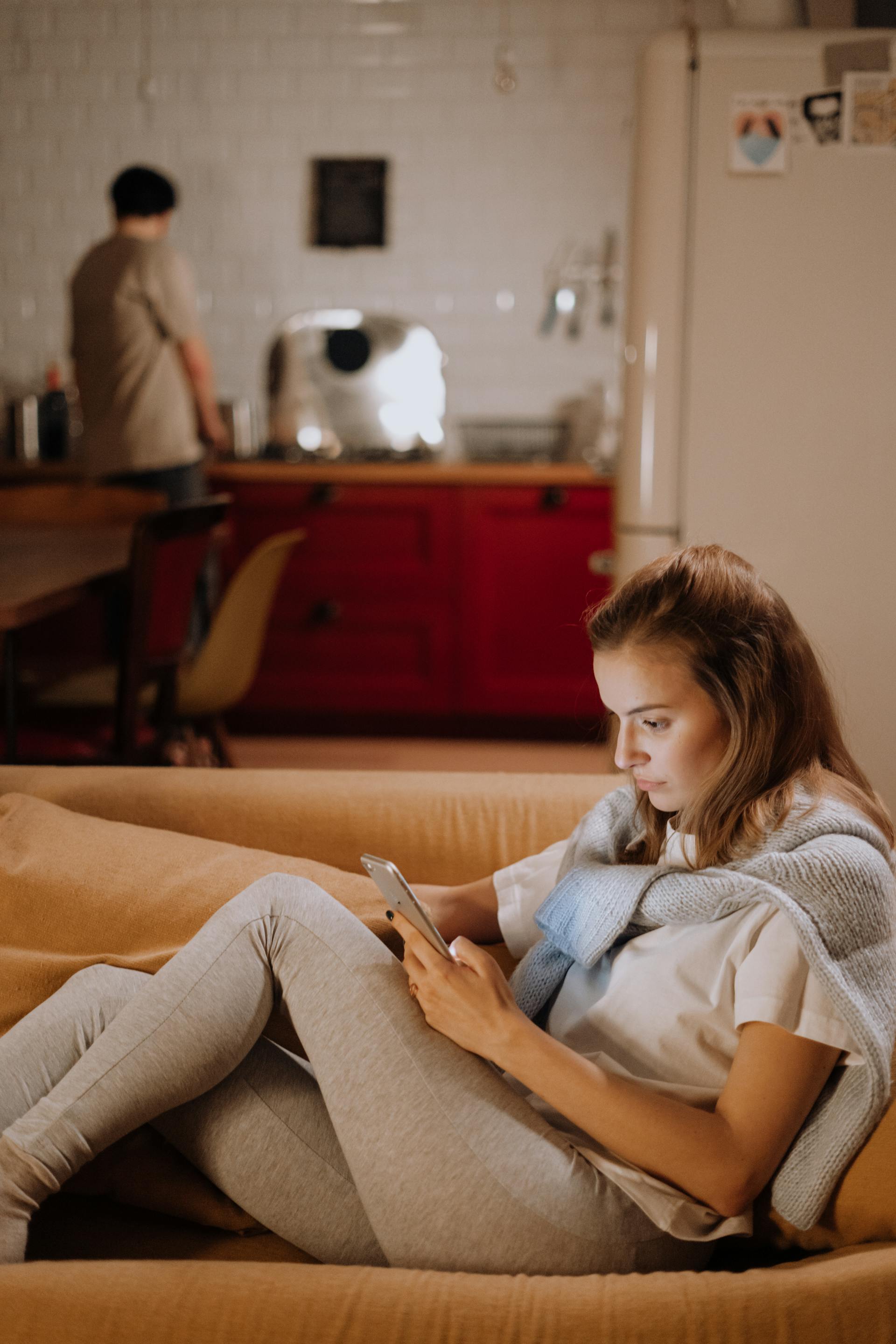 A woman sitting on the couch and using her phone while a man is standing in the kitchen | Source: Pexels