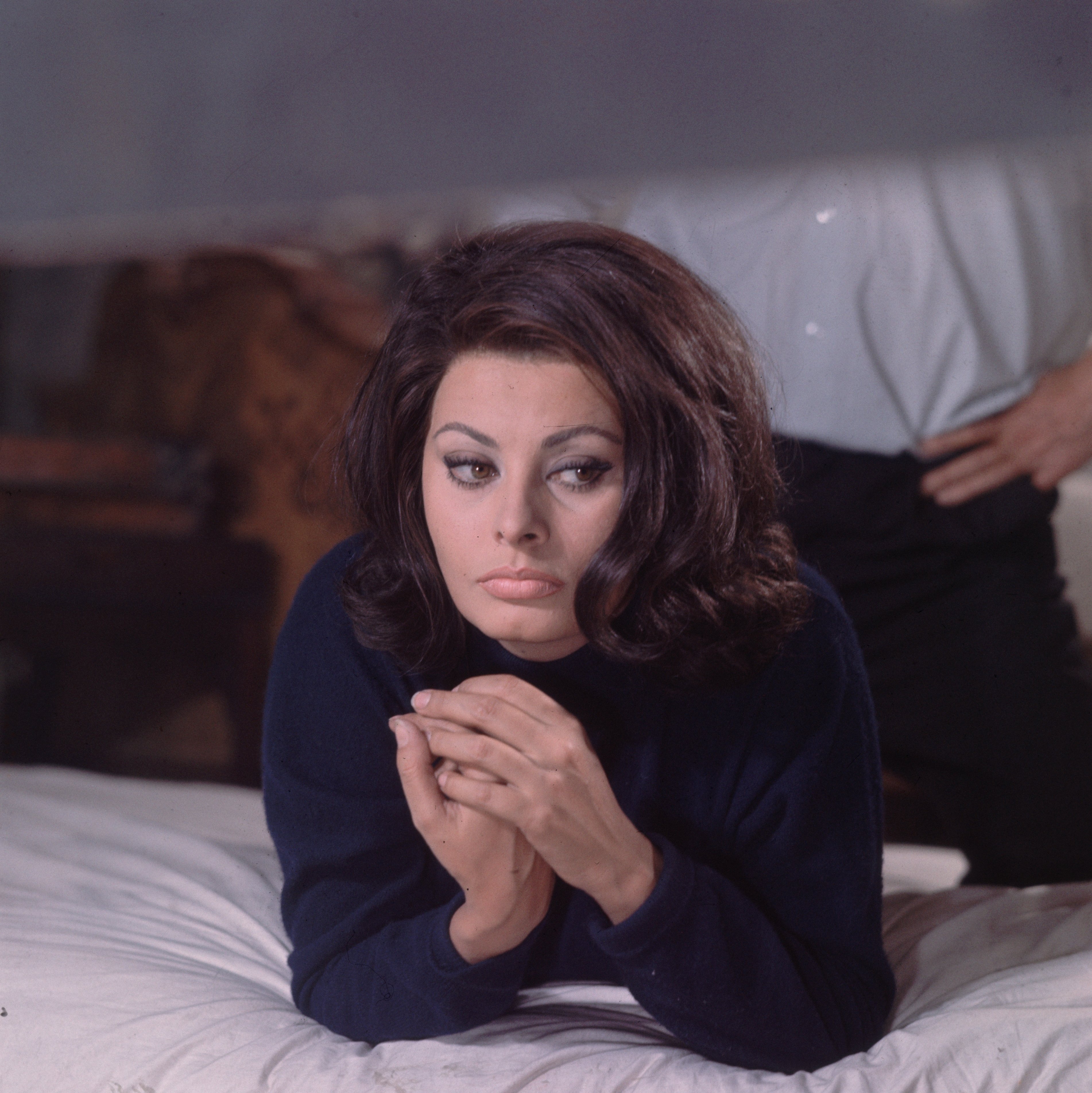 Sophia Loren liegt in einem blauen Rollkragenpullover auf einem Bett, während ein unbekannter Mann im November 1964 in London, England, hinter ihr steht. | Quelle: Getty Images