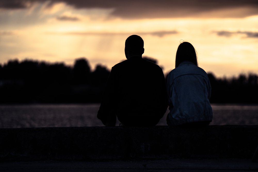 A silhouetted couple sitting next to each other to watch the sunset | Photo: Shutterstock/OlegRi