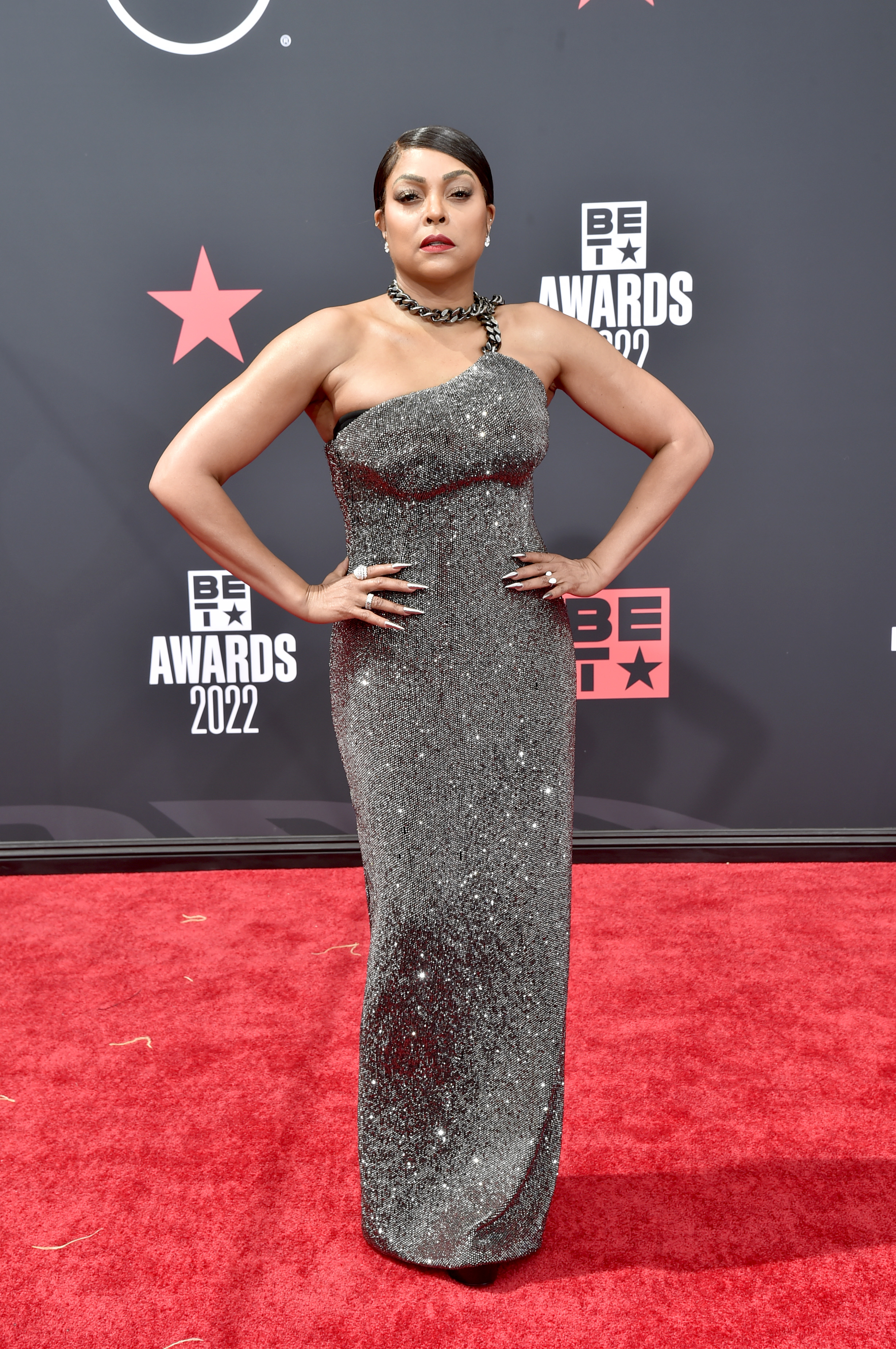 Taraji P. Henson attends the BET Awards at Microsoft Theater in a one-shoulder, metallic-textured gown with a chain-style strap, planting both hands firmly on her hips while standing squarely toward the cameras.