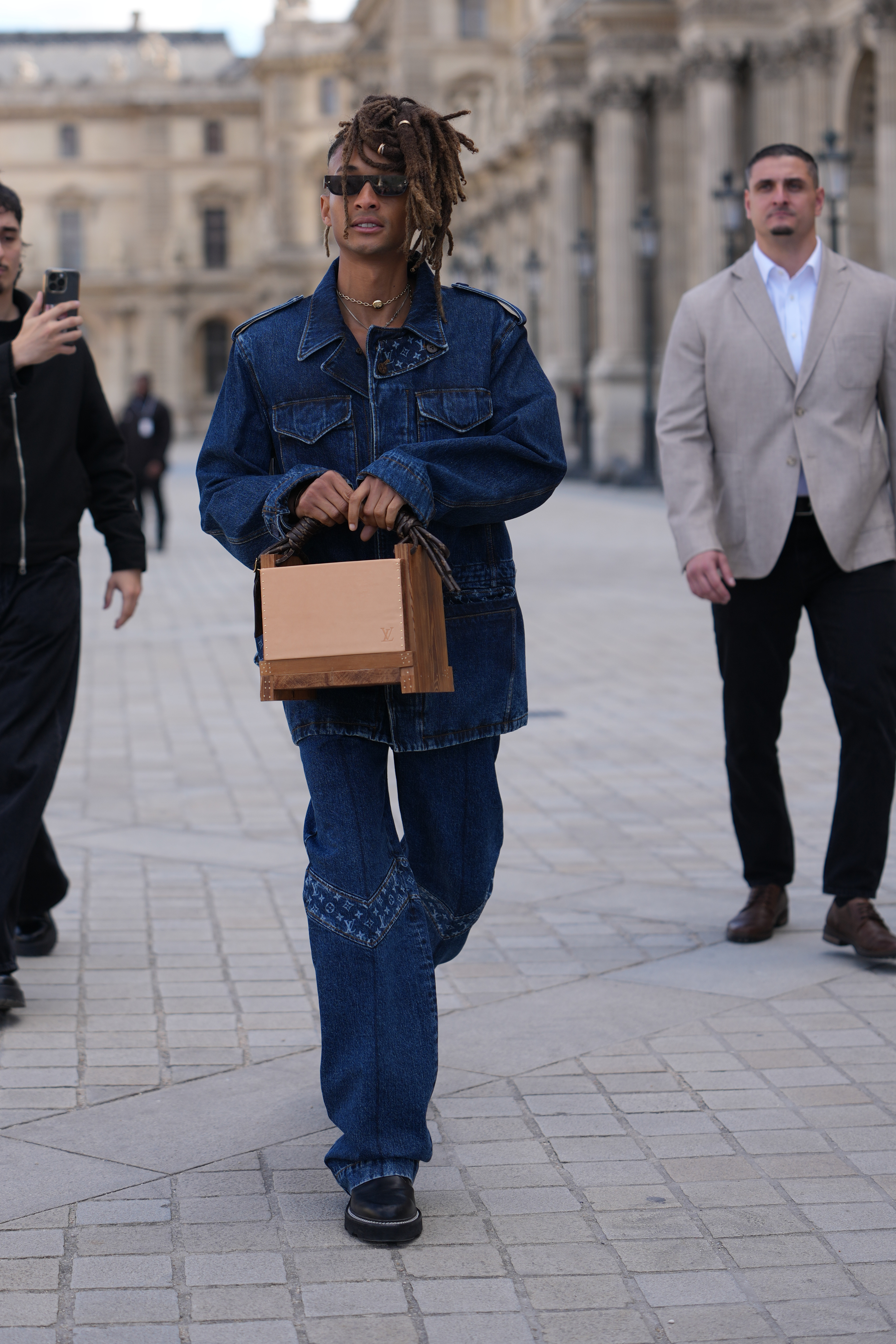 Jaden Smith was spotted outside Louis Vuitton during Paris Fashion Week Women’s Spring/Summer 2026 in France, wearing black rectangular sunglasses and a gold chain necklace. He donned a dark indigo denim jacket, paired with matching relaxed straight-leg jeans featuring V-shaped knee panels. He completed the look with a wooden box clutch and black leather platform slip-on shoes with a low block heel.