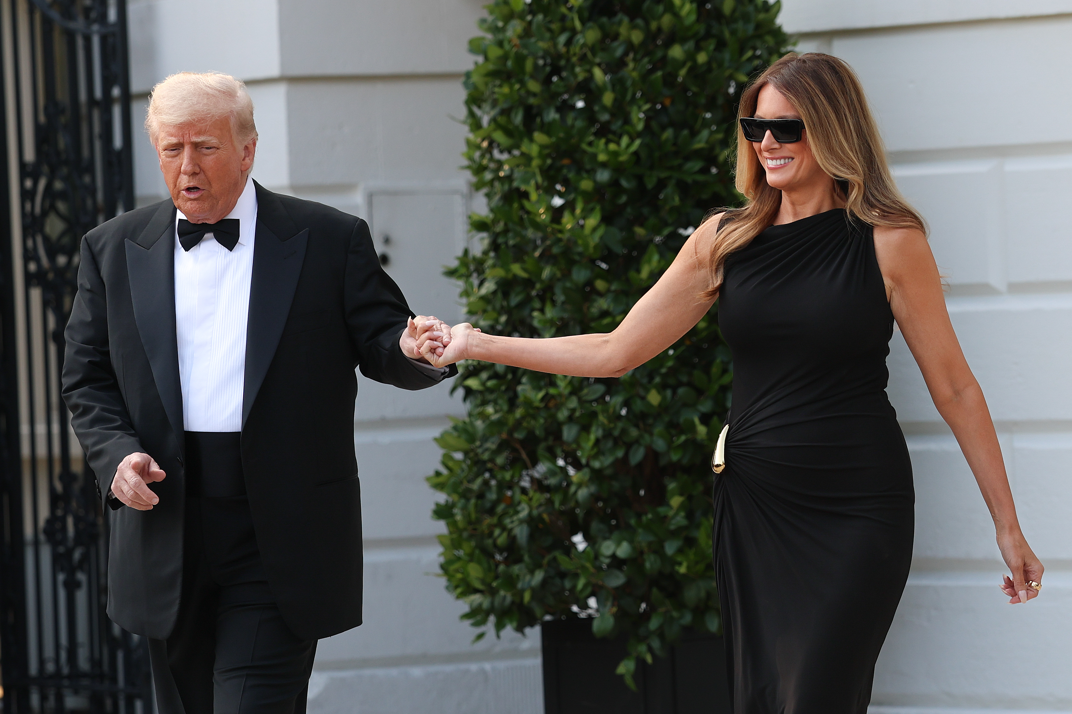 Donald and Melania walk hand in hand, both mid-step and sharing a light moment. Melania smiles brightly behind dark sunglasses, the curve of her black gown more visible as it gathers at the hip with a gold clasp.