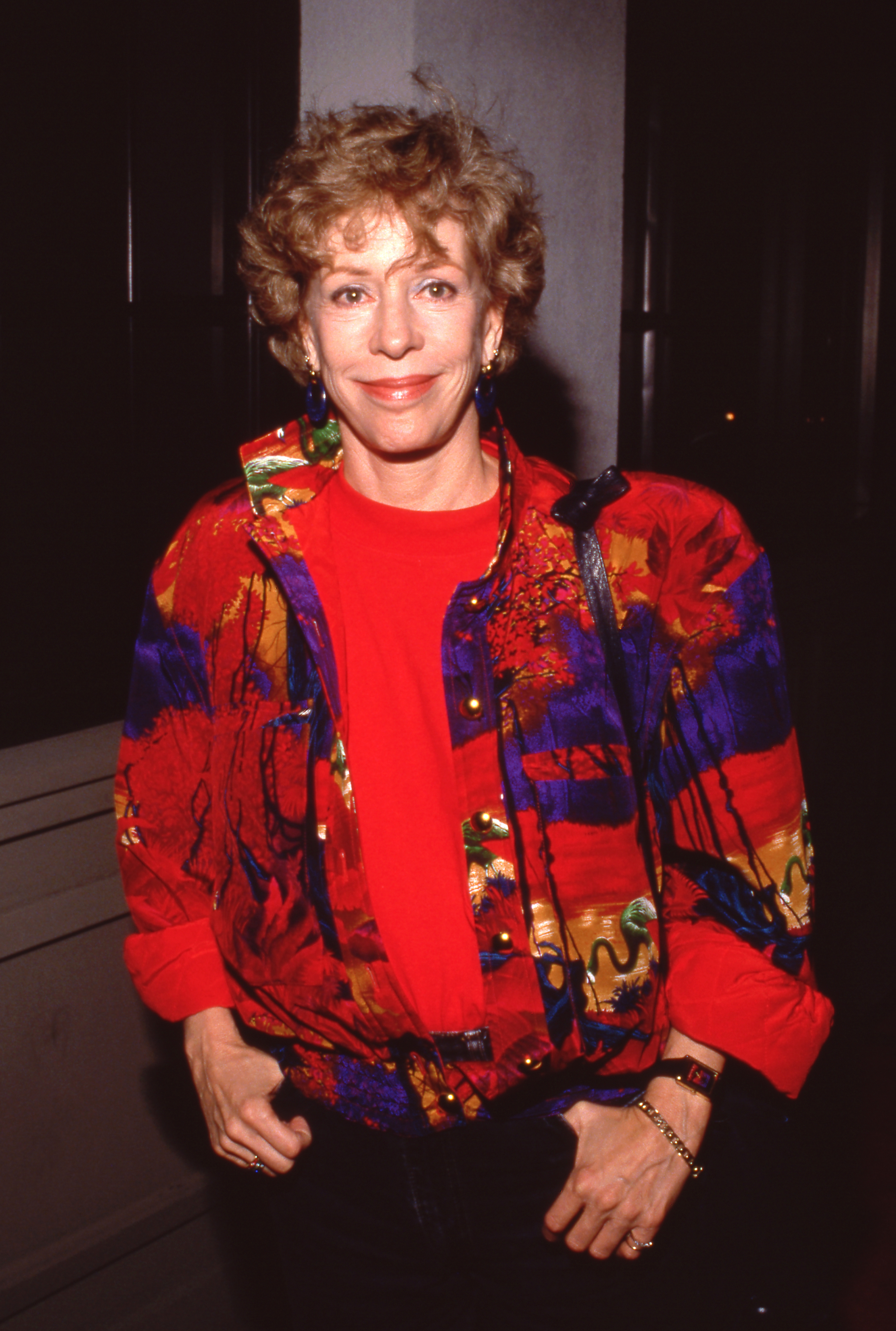 She arrives in a fitted black dress layered with a red-and-gold patterned jacket. The jacket features intricate detailing, and she stands with her hands together at waist level.
