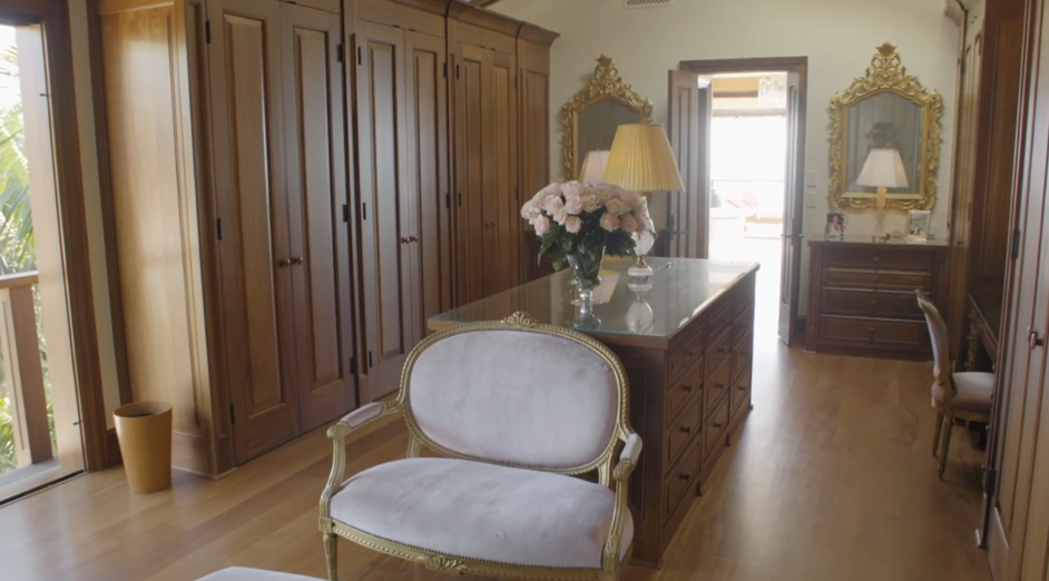 Rich warm wood cabinetry encloses the extensive primary suite closet, providing ample storage behind seamless panel doors. A glass-topped center island topped with a bouquet of pale pink roses sits between ornate gold-framed mirrors, a vanity seating area, and an opening that leads directly toward the bright bedroom space. | Source: YouTube/Architectural Digest