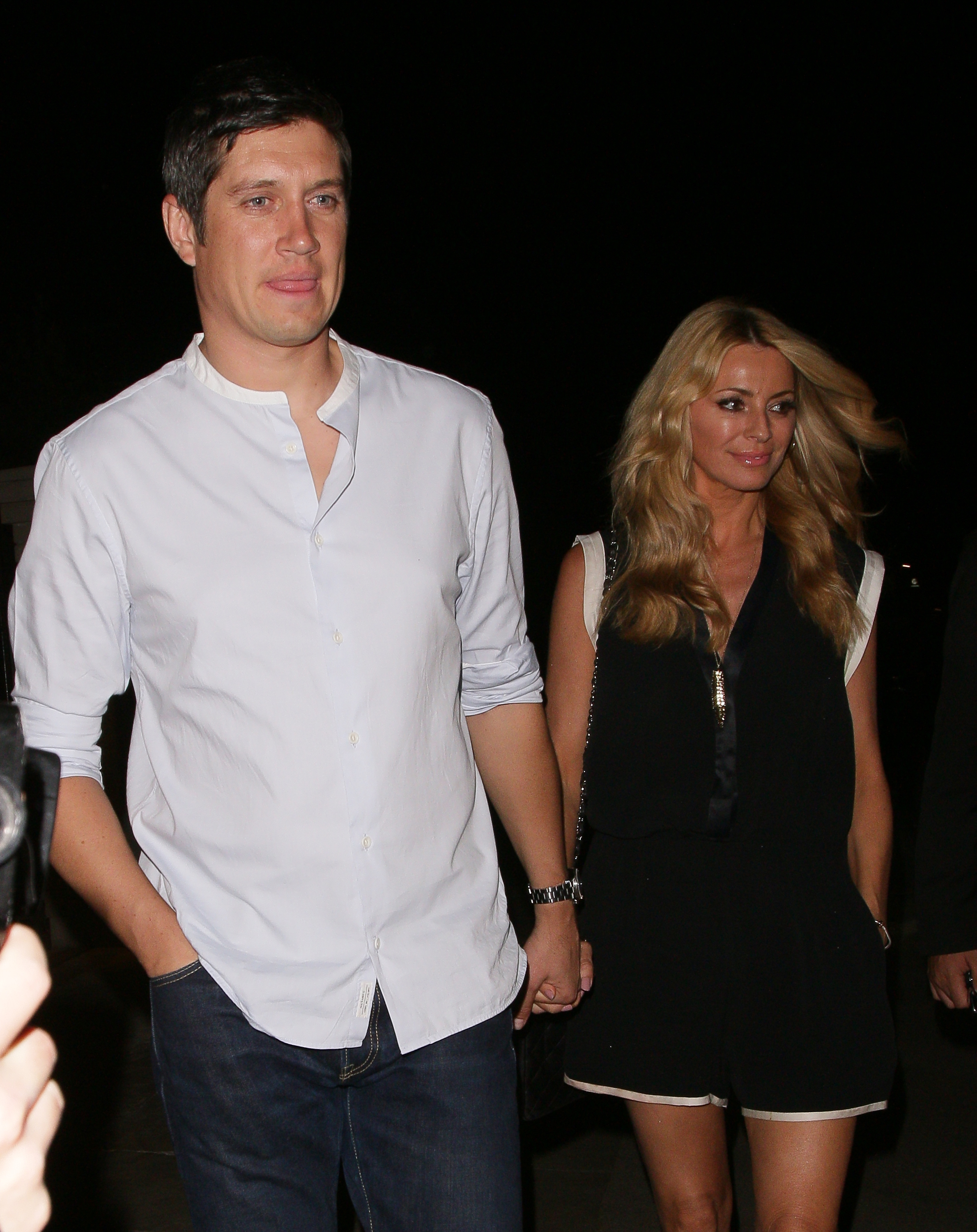 Vernon Kay, in a light button-down shirt and jeans, walks hand in hand with Tess Daly, who wears a sleeveless black outfit, as the pair step out together at night.