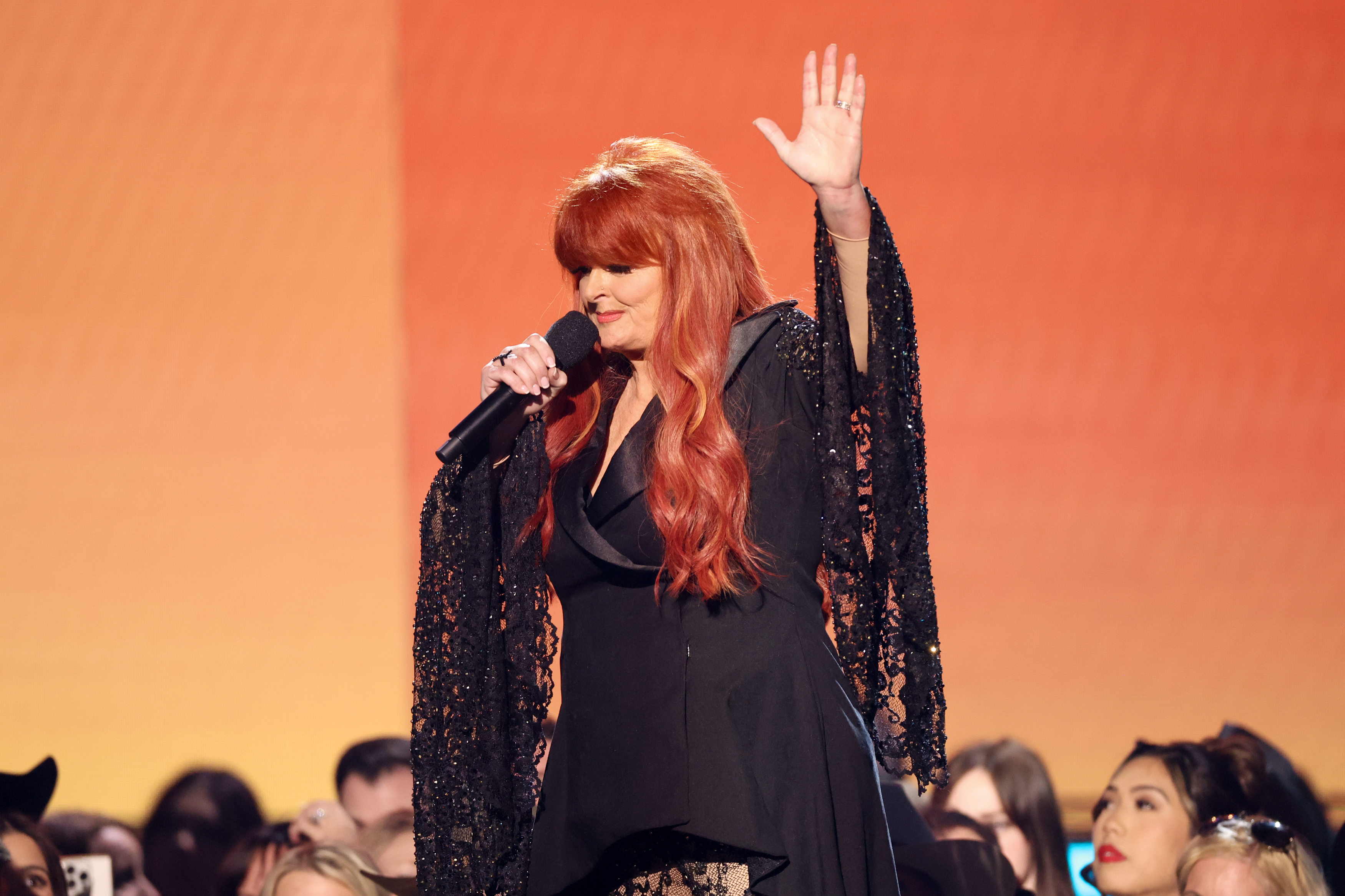 Wynonna Judd is seen onstage at the 60th ACM Awards at the Ford Center in Frisco, Texas. With one hand raised and a mic in the other, she addresses the crowd, her glittering sleeves catching the light as fans look on from the audience.