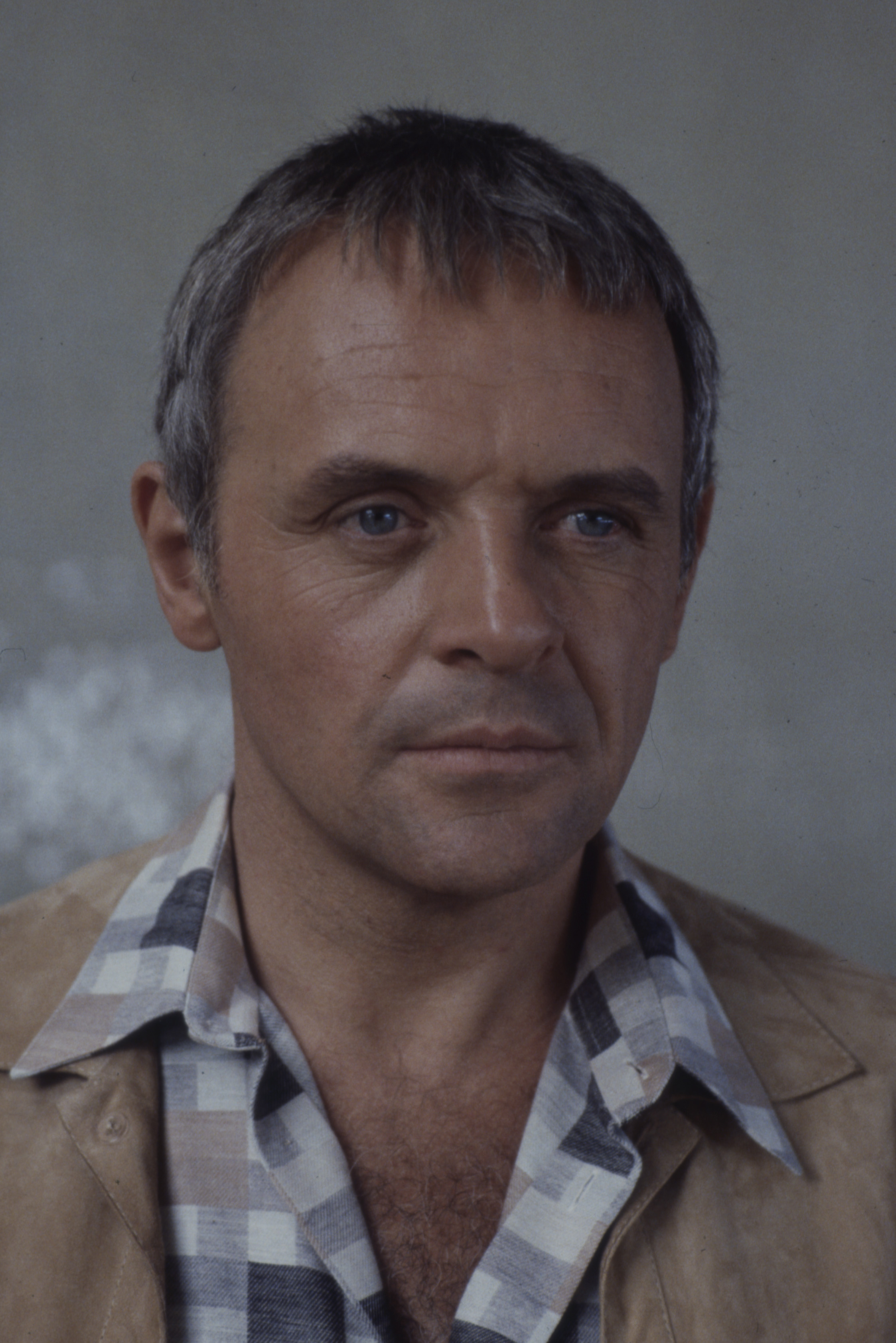 Appearing contemplative and composed, Anthony Hopkins's piercing blue eyes reflects quiet intensity. Dressed in a checkered shirt and tan jacket, he embodies understated class. The muted tones and soft lighting capture a moment of depth and quiet drama from "Hollywood Wives."