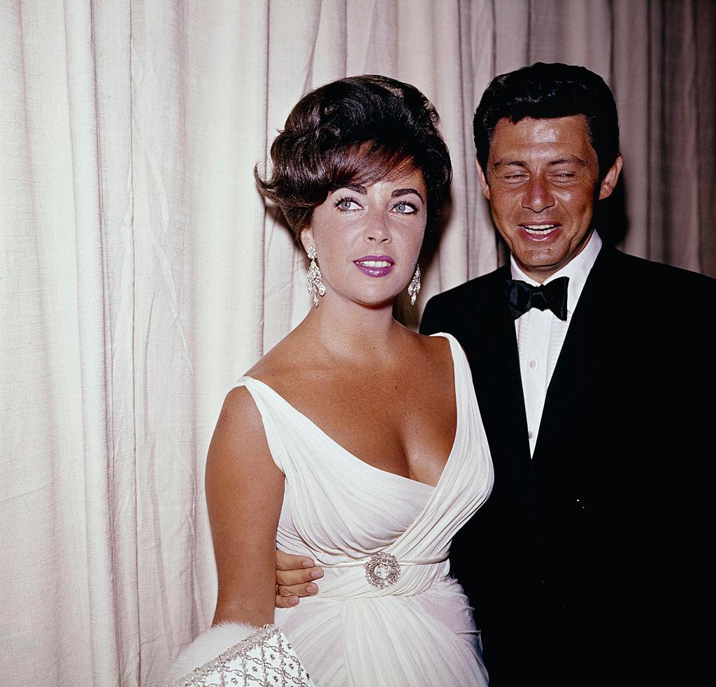 Elizabeth Taylor und Eddie Fisher nehmen an einer Veranstaltung teil, ca. 1950-60. Taylor trägt ein weißes Kleid mit einer Brosche in der Taille. | Quelle: Getty Images