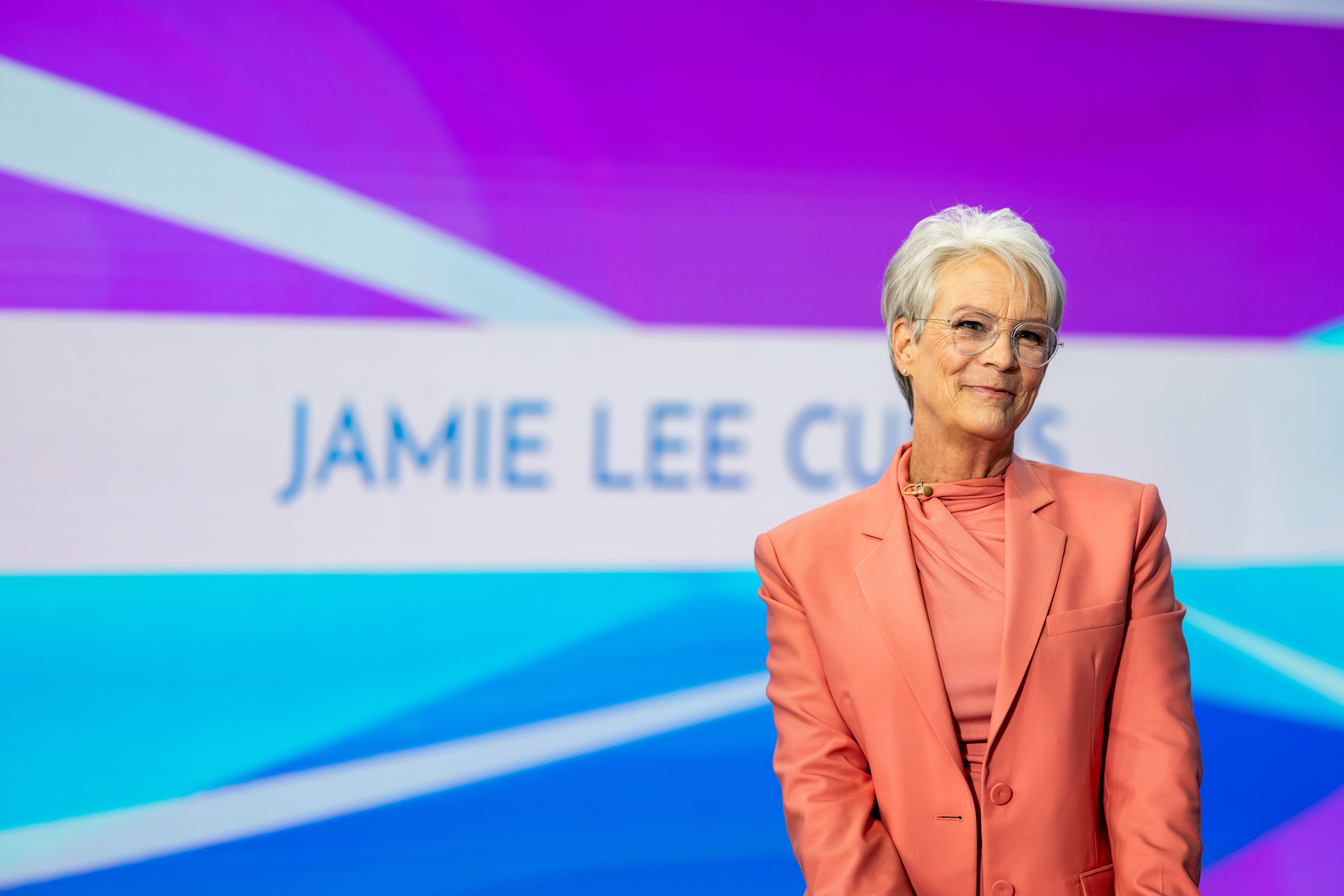 Jamie Lee Curtis appears on “TODAY” in a vibrant coral suit and matching draped blouse, her silver hair styled in a soft crop as she smiles confidently against a bold, colorful backdrop.