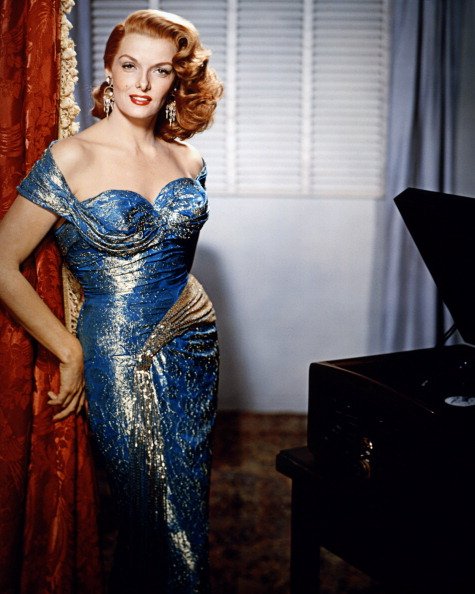 Photo of Jane Russell wearing an off-the-shoulder blue and gold dress, with a red theatre curtain behind her, circa 1955 | Photo: Getty Images