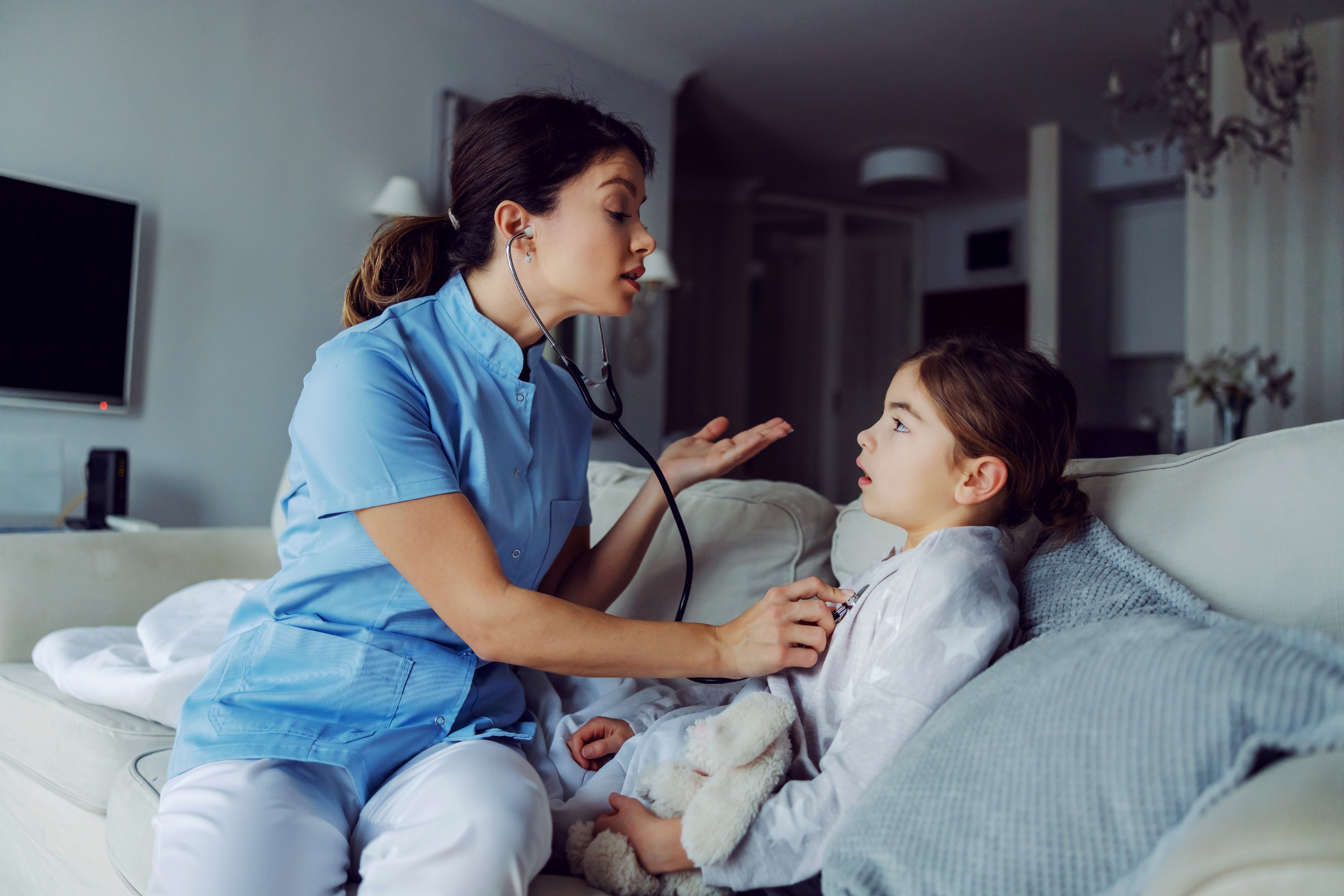 Doctor sitting on sofa next to girl and examining her lungs with stethoscope | Source: Shutterstock.com