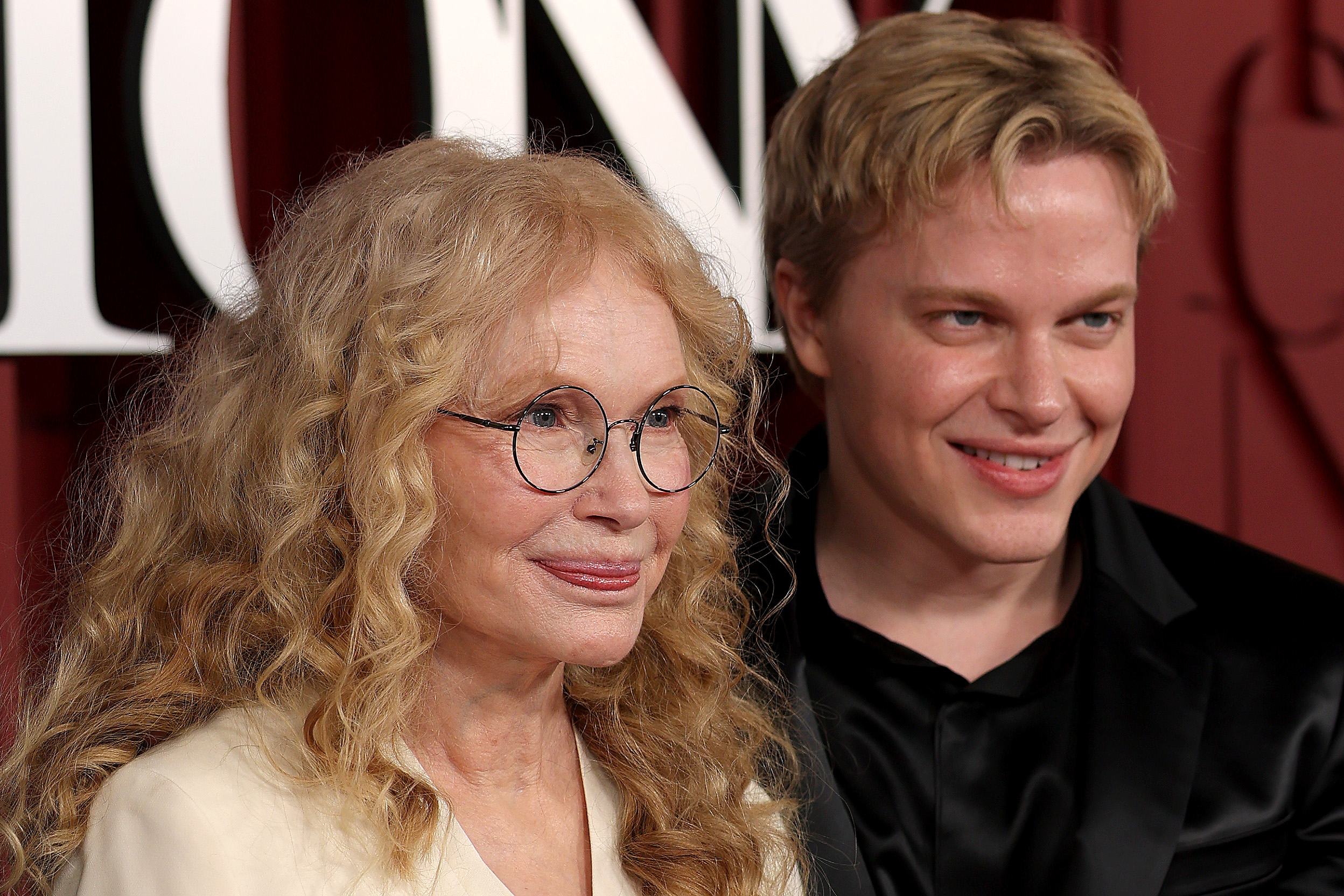 Mia Farrow and Ronan Farrow pose close together, smiling softly under the lights. Her iconic curls and glasses meet his polished look and subtle smirk, capturing a tender moment that feels both timeless and full of quiet charm.