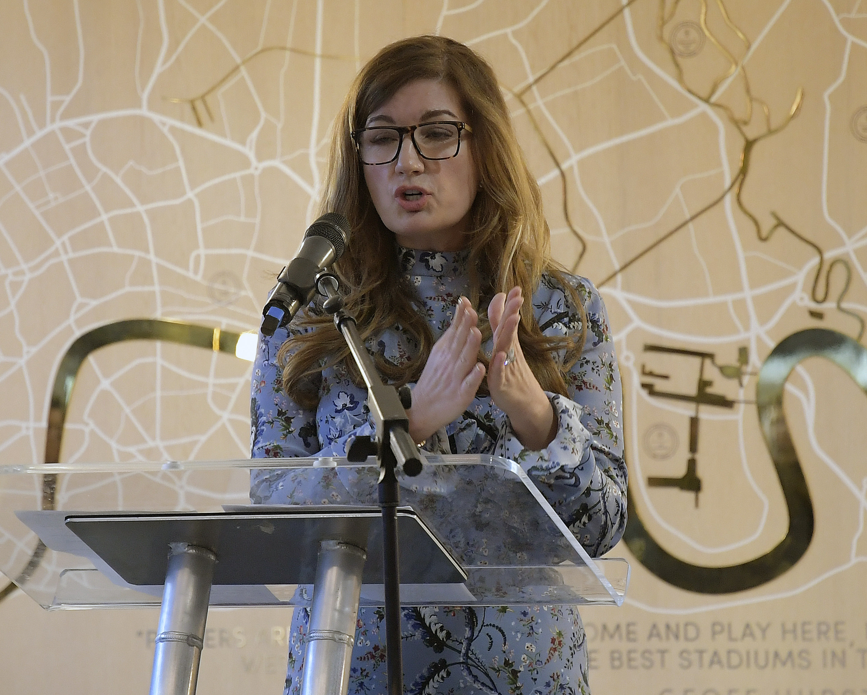 Commanding the Room: Baroness Brady stands tall at the podium during The Players’ Project launch on 7 November 2018, addressing the media from London Stadium. Wearing a patterned blue dress and glasses, she exudes gravitas and grace, as she outlines a vision to inspire change through sport.