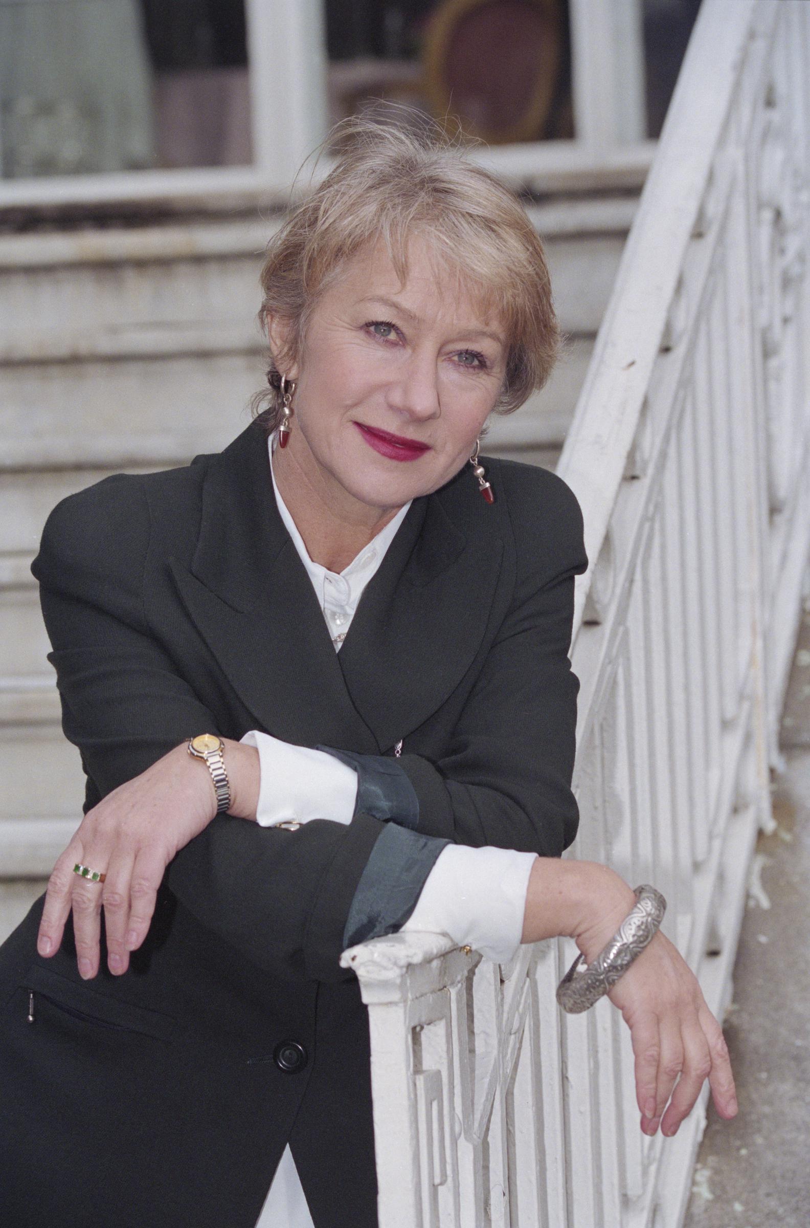 Helen Mirren leans gracefully against a white railing in this poised outdoor portrait, dressed in a dark structured blazer with contrasting cuffs, her look completed by bold red lipstick, drop earrings, and a statement bangle that adds a touch of character to her polished ensemble.