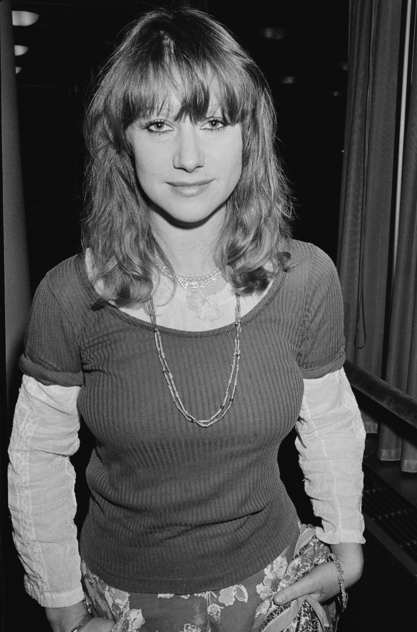 A young Helen Mirren poses for a candid black-and-white portrait in the 1970s, wearing layered necklaces and a ribbed top over a patterned skirt, her relaxed expression framed by tousled hair and curtain bangs.