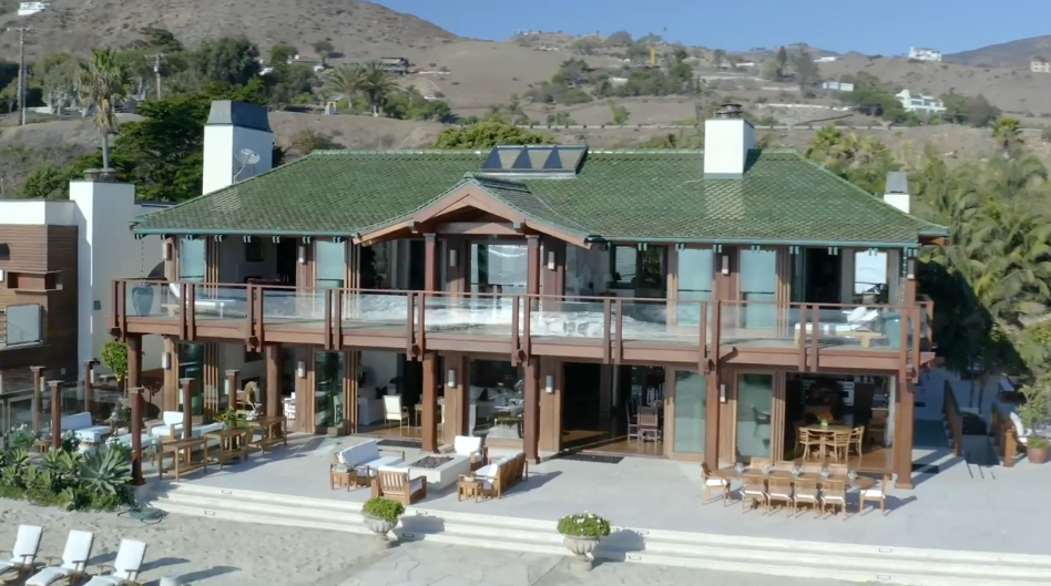 An elevated view captures the full rear exterior of Orchid House, showcasing its distinctive green clay-tiled roof, rich timber columns, and sweeping second-story glass balcony. Wide steps descend from the main terrace—furnished with multiple lounge setups and a long dining table—directly onto the sandy beach against the backdrop of the Malibu hills. | Source: YouTube/Architectural Digest