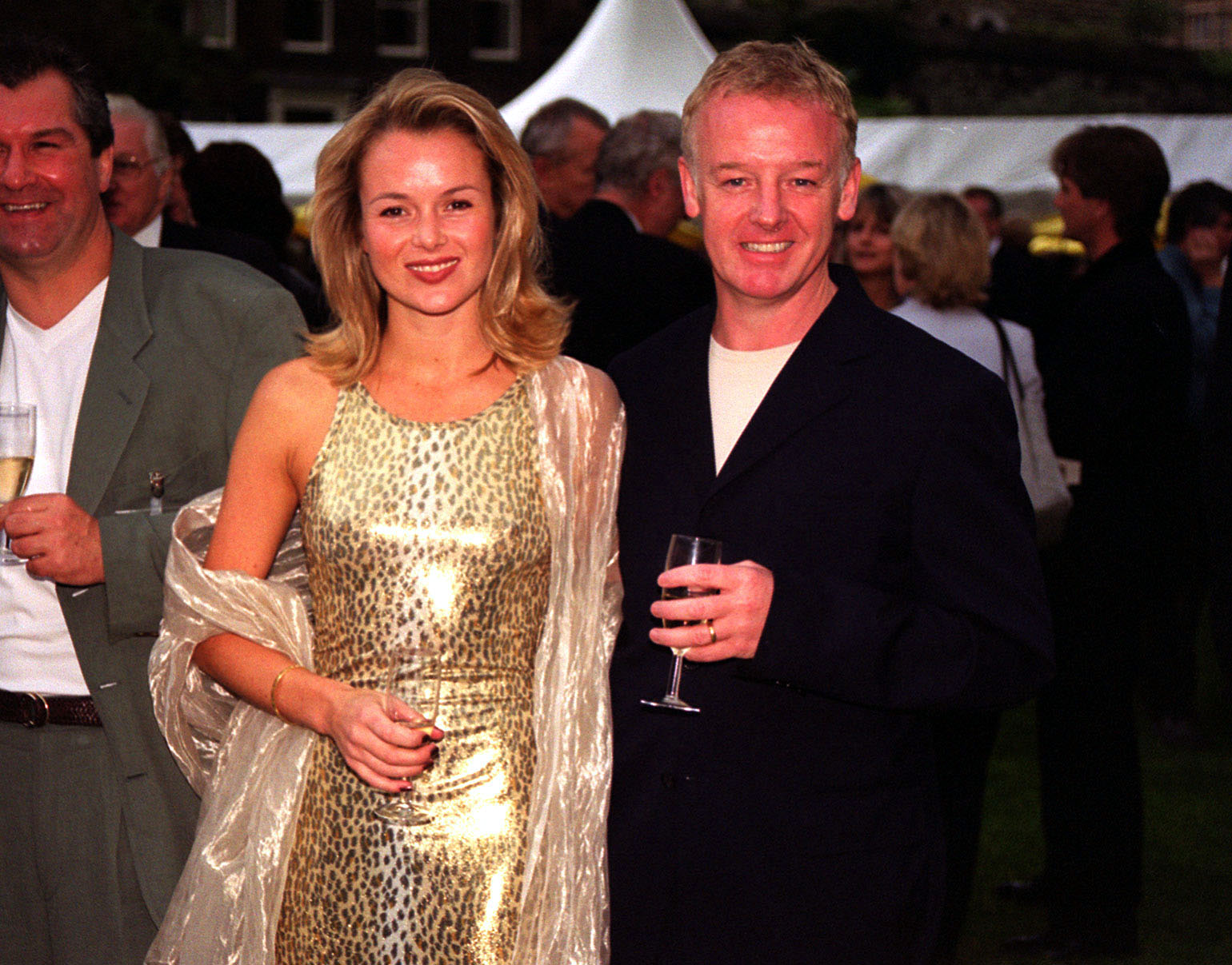 Holden, in a gold leopard-print slip dress with a sheer wrap, and Dennis, in a black blazer and white T-shirt, hold champagne flutes at the Carlton Television Parliamentary Summer reception in the grounds of Westminster Abbey.