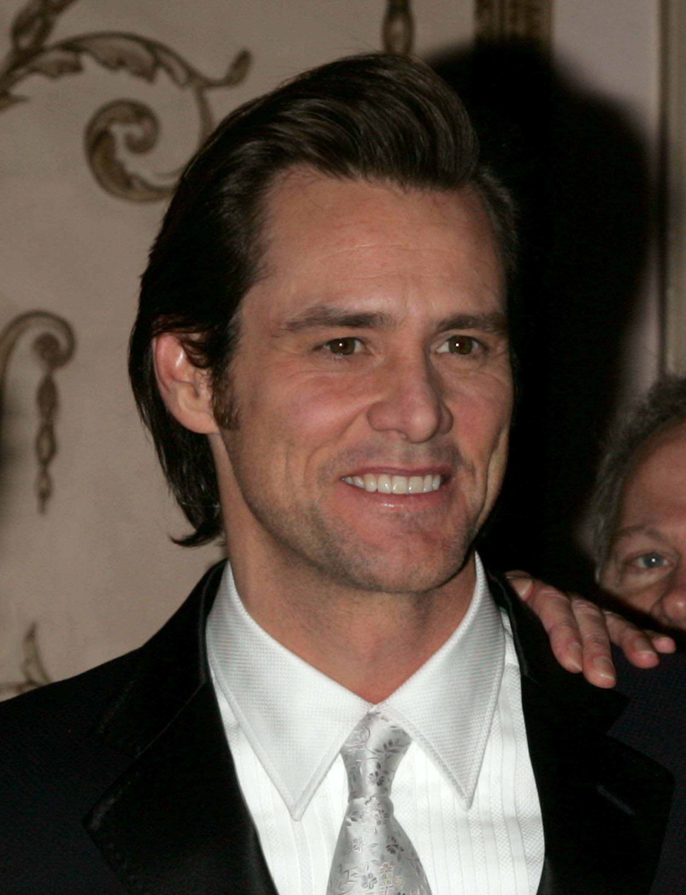 Jim Carrey is captured at the Museum of the Moving Image Salute to Ron Howard, dressed elegantly in a black suit with a white shirt and patterned silver tie. He is smiling warmly, exuding confidence and charm in the formal setting. The event’s refined atmosphere complements his polished and charismatic appearance.