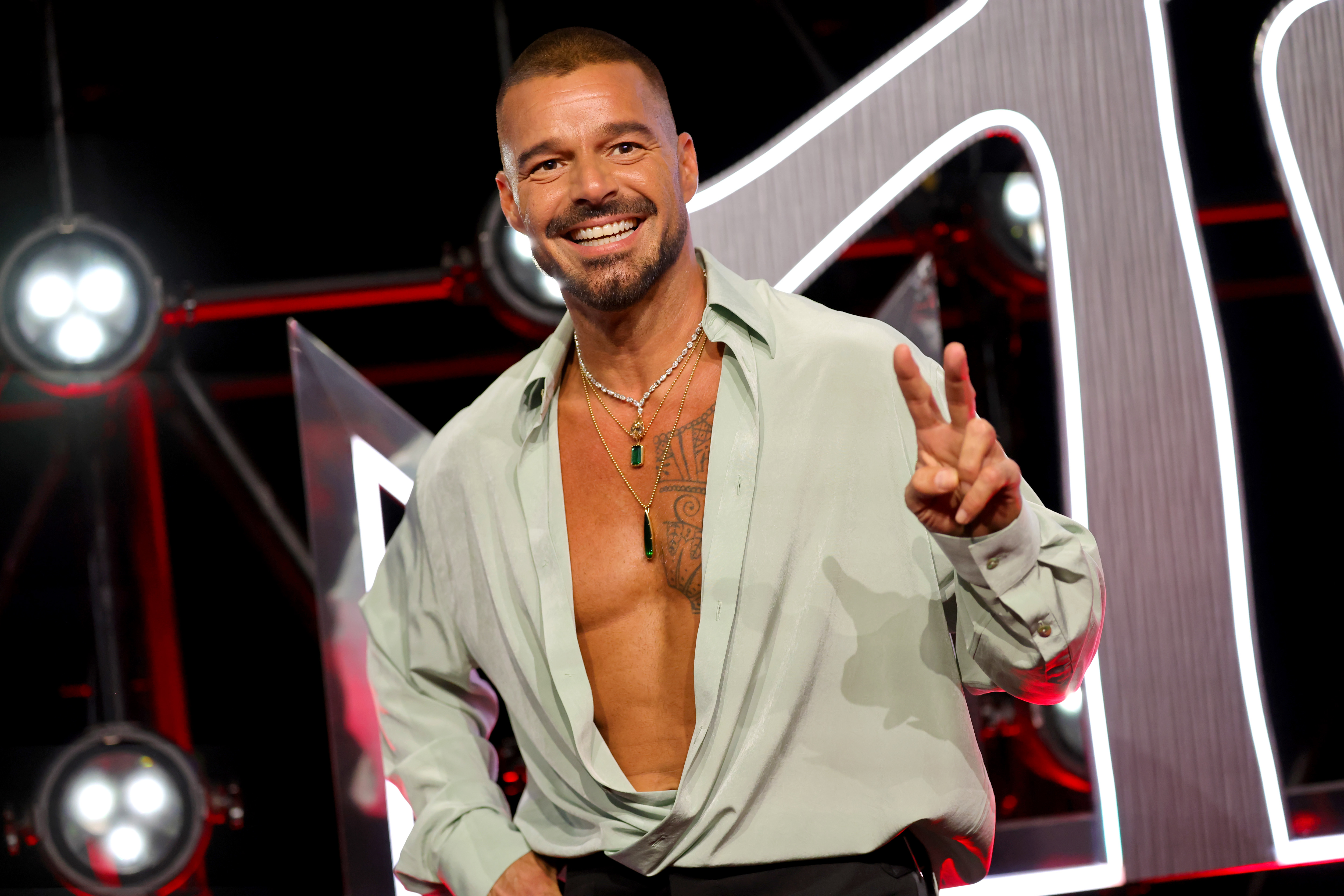 Ricky Martin flashes a wide smile and raises a peace sign while wearing an open pale green shirt and layered necklaces at the 2025 MTV Video Music Awards in Elmont, New York.