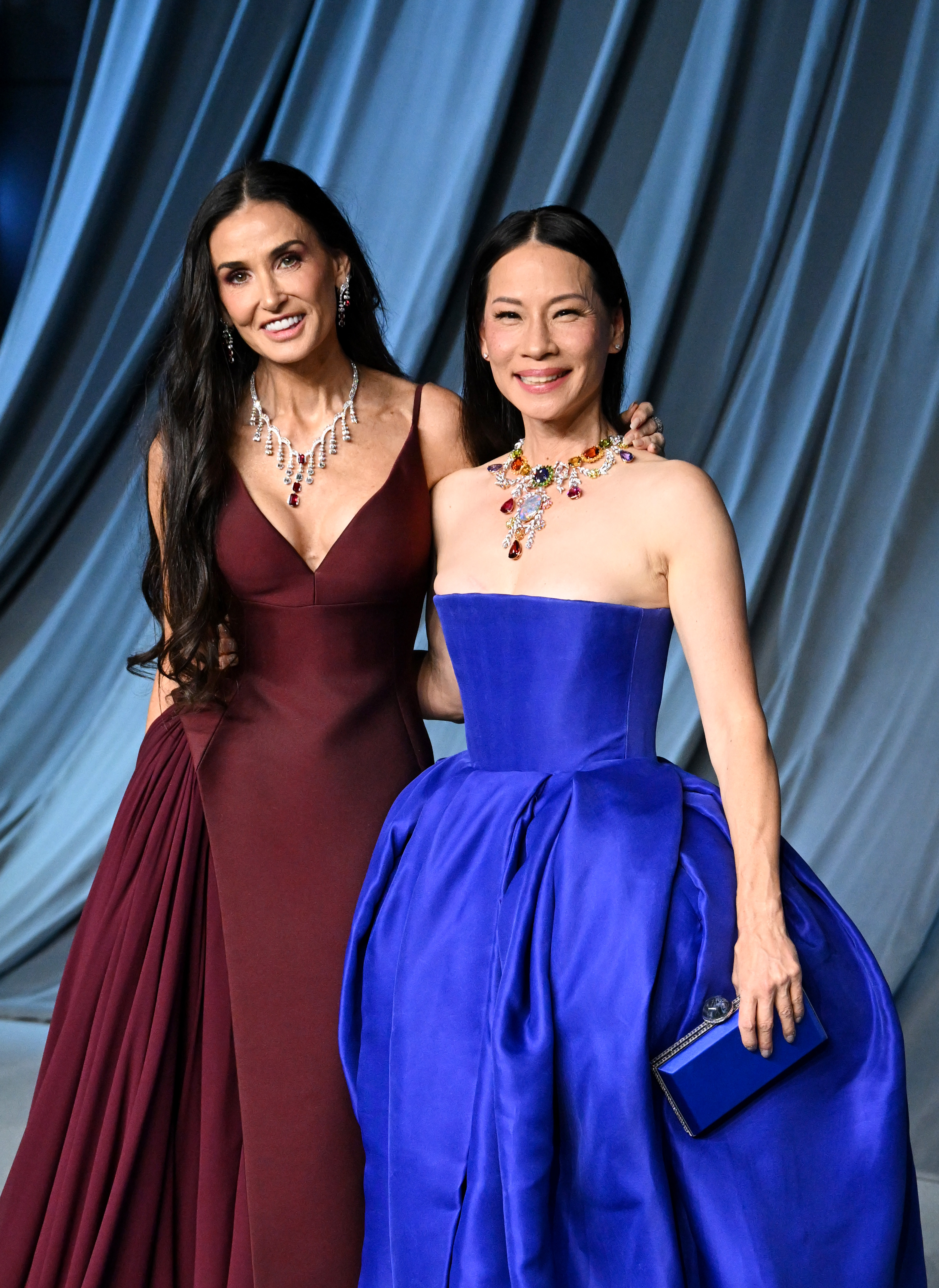 Smiling effortlessly beside Lucy Liu, Demi Moore radiates relaxed glamour in her curve-skimming maroon gown, while Liu stuns in royal blue with a vibrant jeweled neckline — a true power duo moment.