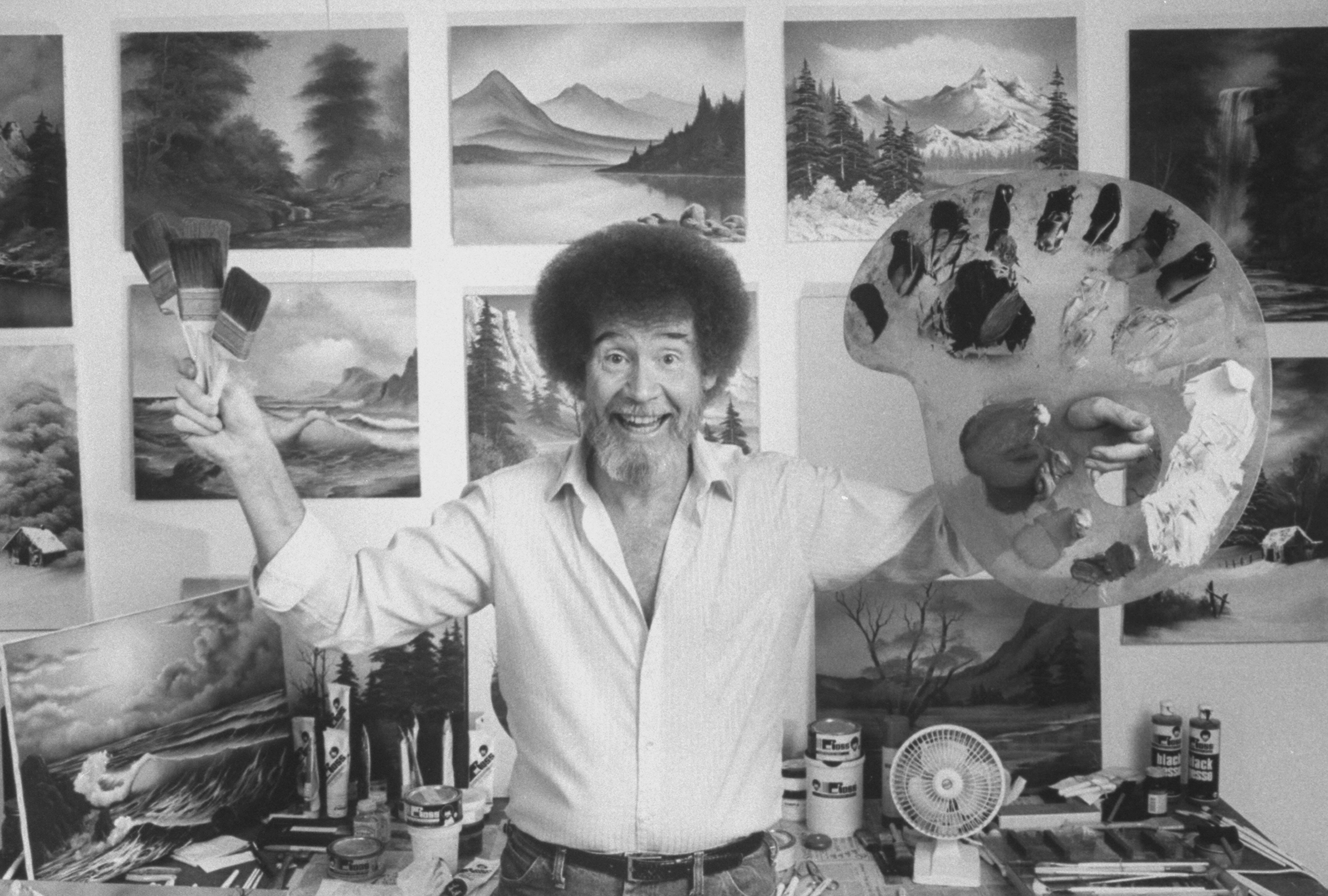Bob Ross holding up a paint palette & brushes as he stands in front of a wall covered with his landscape paintings in his studio in 1991. | Source: Getty Images