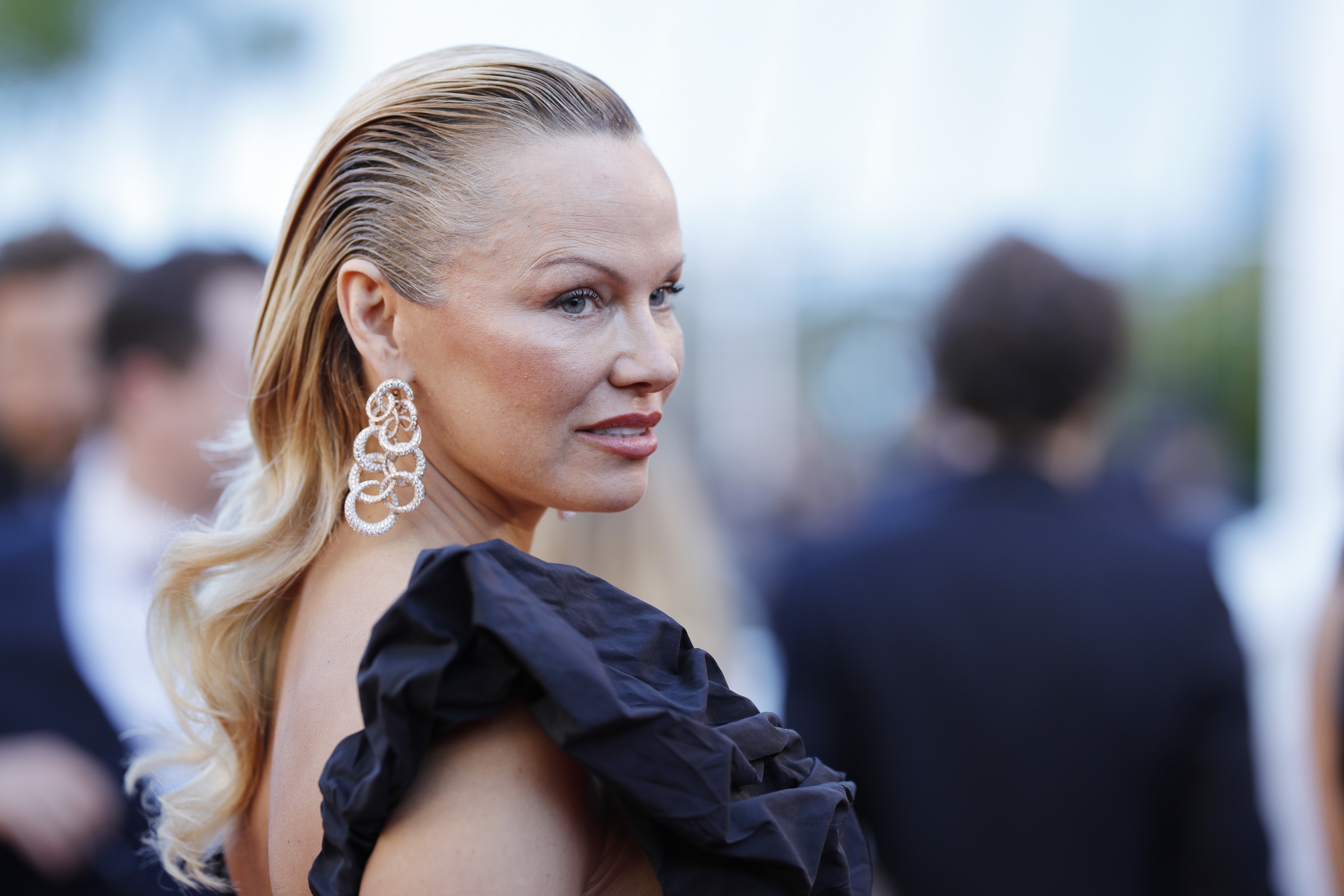 Pamela Anderson exudes elegance in a sleek black dress, her long blonde hair styled back for a polished, refined look. Gold earrings add a touch of sophistication, subtly highlighting her features and completing the timeless ensemble with understated glamour.