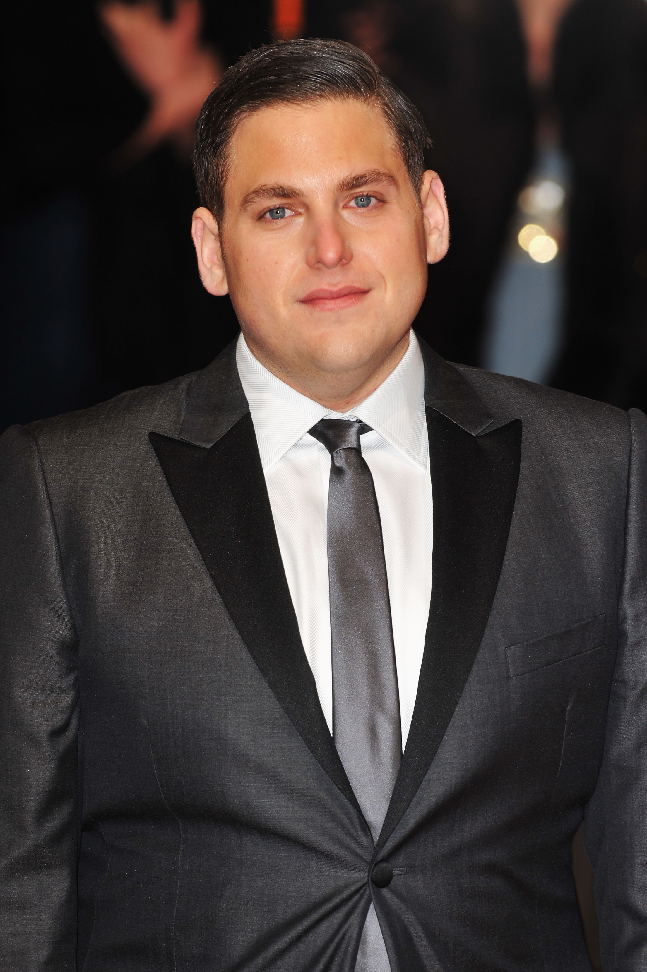 Jonah Hill steps out on the red carpet in a sharp charcoal tuxedo with black satin lapels, paired with a crisp white shirt and matching grey tie. With his hair neatly slicked back, the actor showcases a noticeably slimmer figure at the formal event.