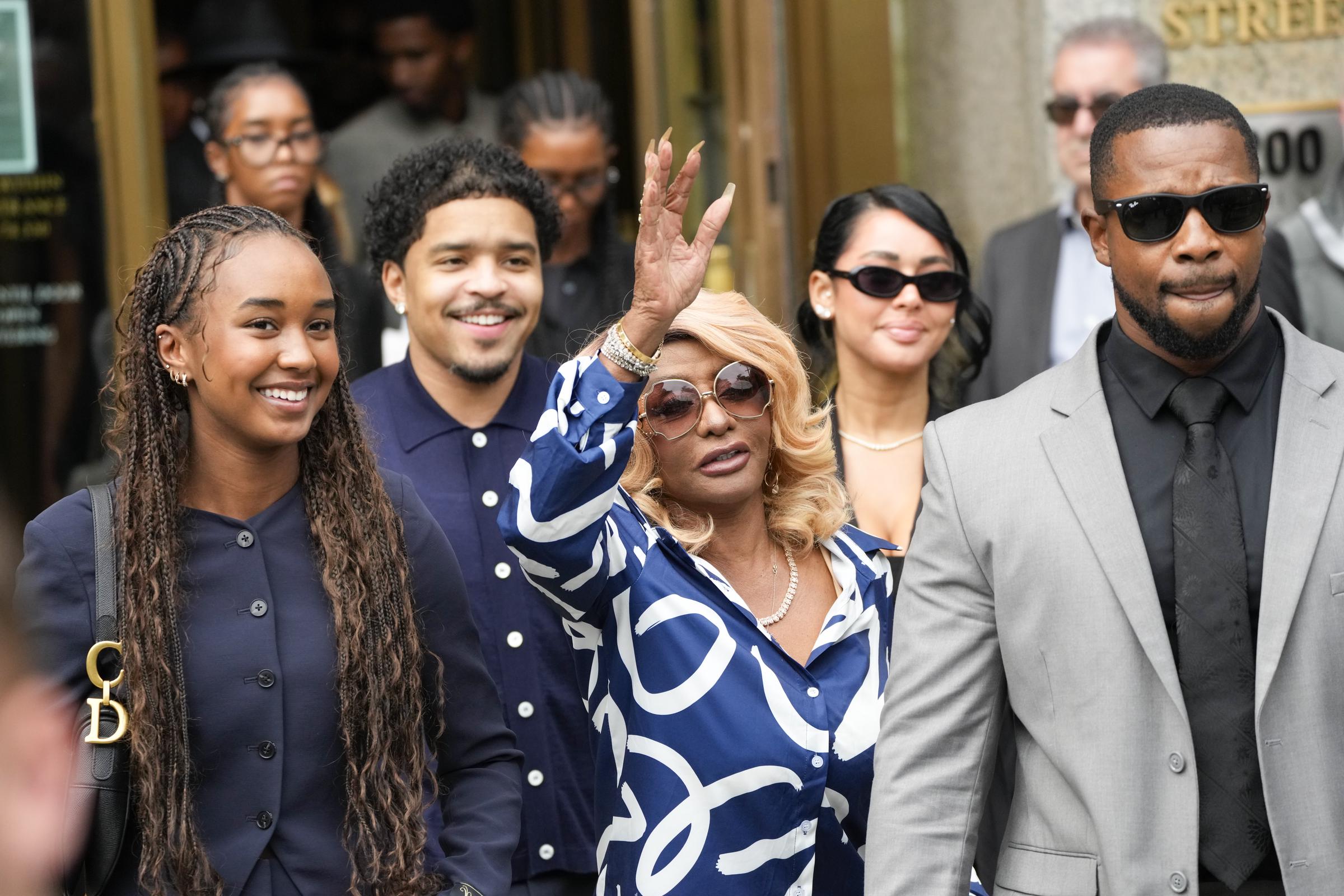 Janice Combs raises a hand in acknowledgment as she leaves the courthouse, flanked by smiling family members. Quincy Brown walks nearby, grinning.