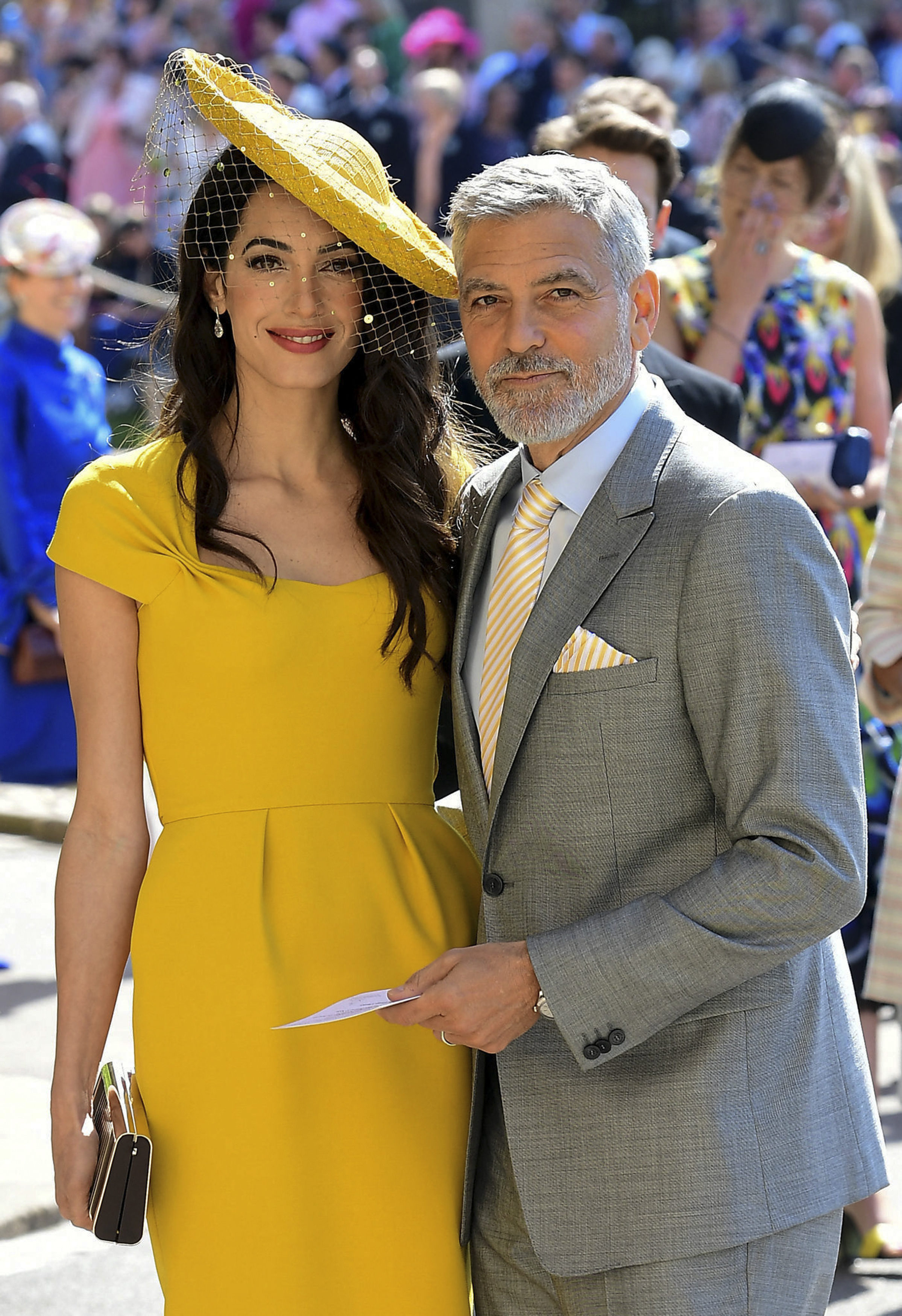 Amal turns heads at the royal wedding of Prince Harry and Meghan Markle in a bright yellow fitted dress with cap sleeves and a matching wide-brimmed hat with a netted veil. She stands beside George, who wears a light gray suit with a yellow-striped tie and pocket square.