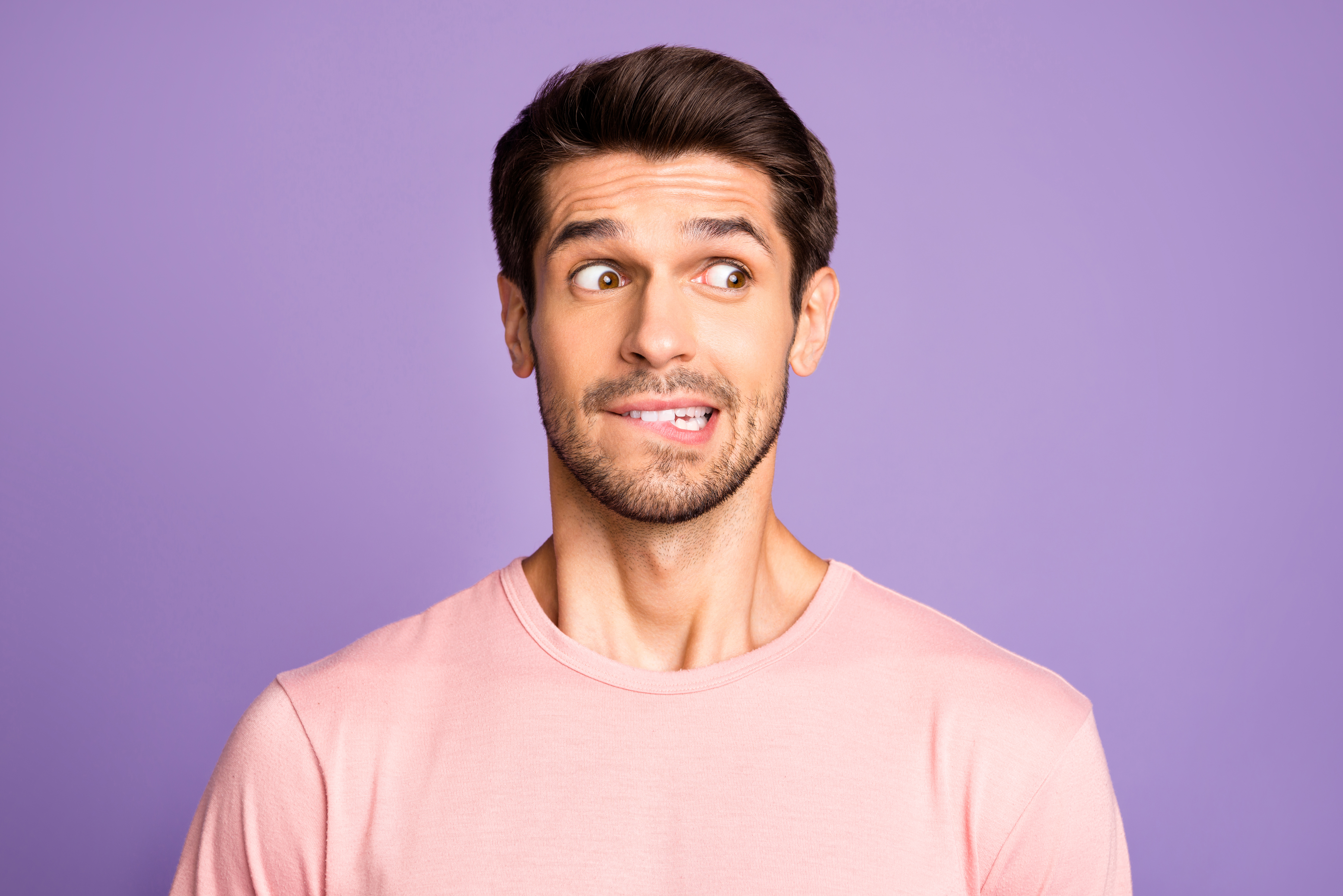 Retrato de primer plano de su chico barbudo moreno, divertido, divertido y divertido, vestido con una camiseta rosa, esperando noticias, mordiéndose el labio aislado sobre un fondo de color pastel lila violeta violeta | Fuente: Getty Images