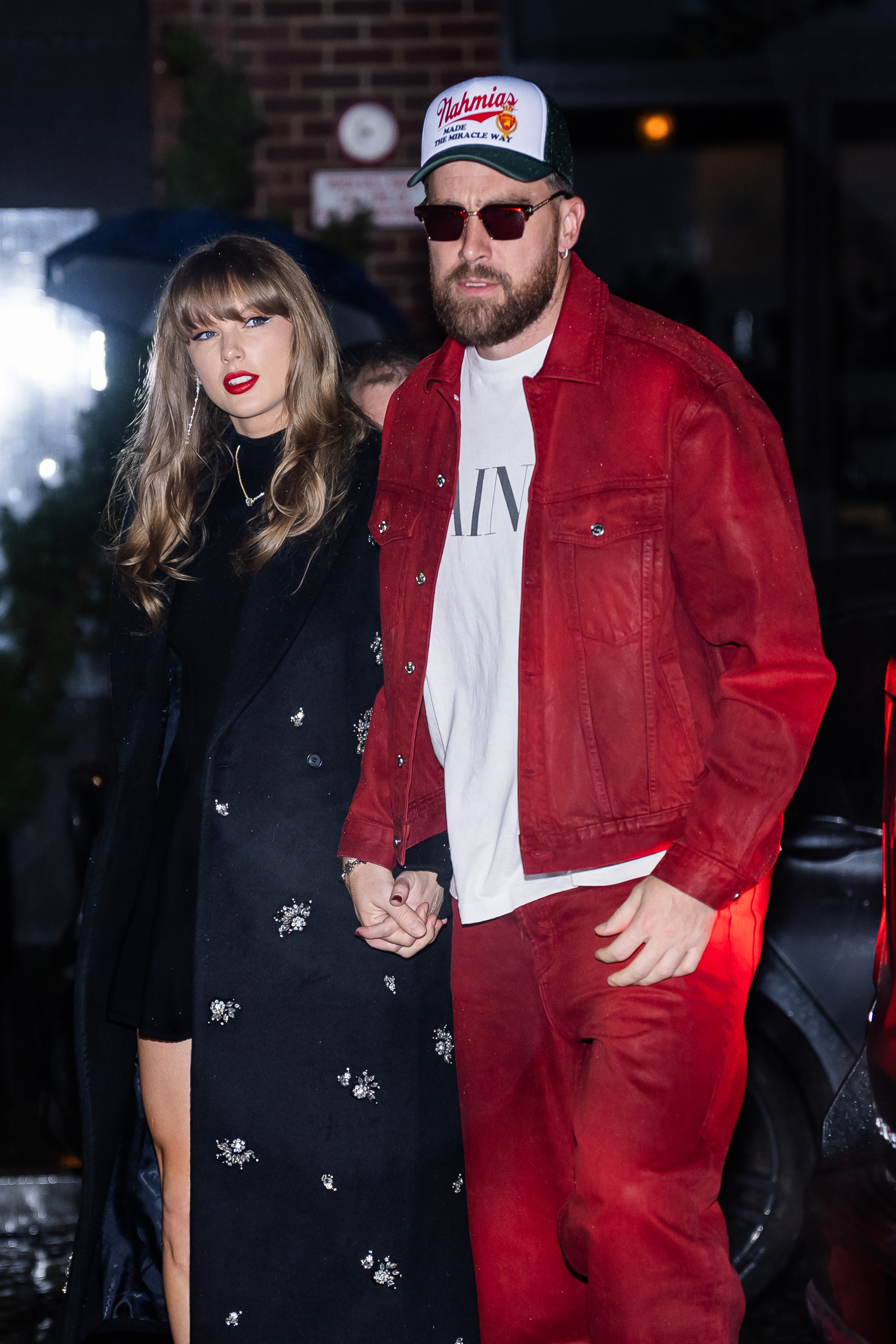 Taylor Swift, wearing a black embellished coat over a mini dress, walks hand in hand with Travis Kelce, dressed in a red jacket and matching pants with a white T-shirt and trucker hat, as they step out in the Meatpacking District on December 28, 2024, in New York City.