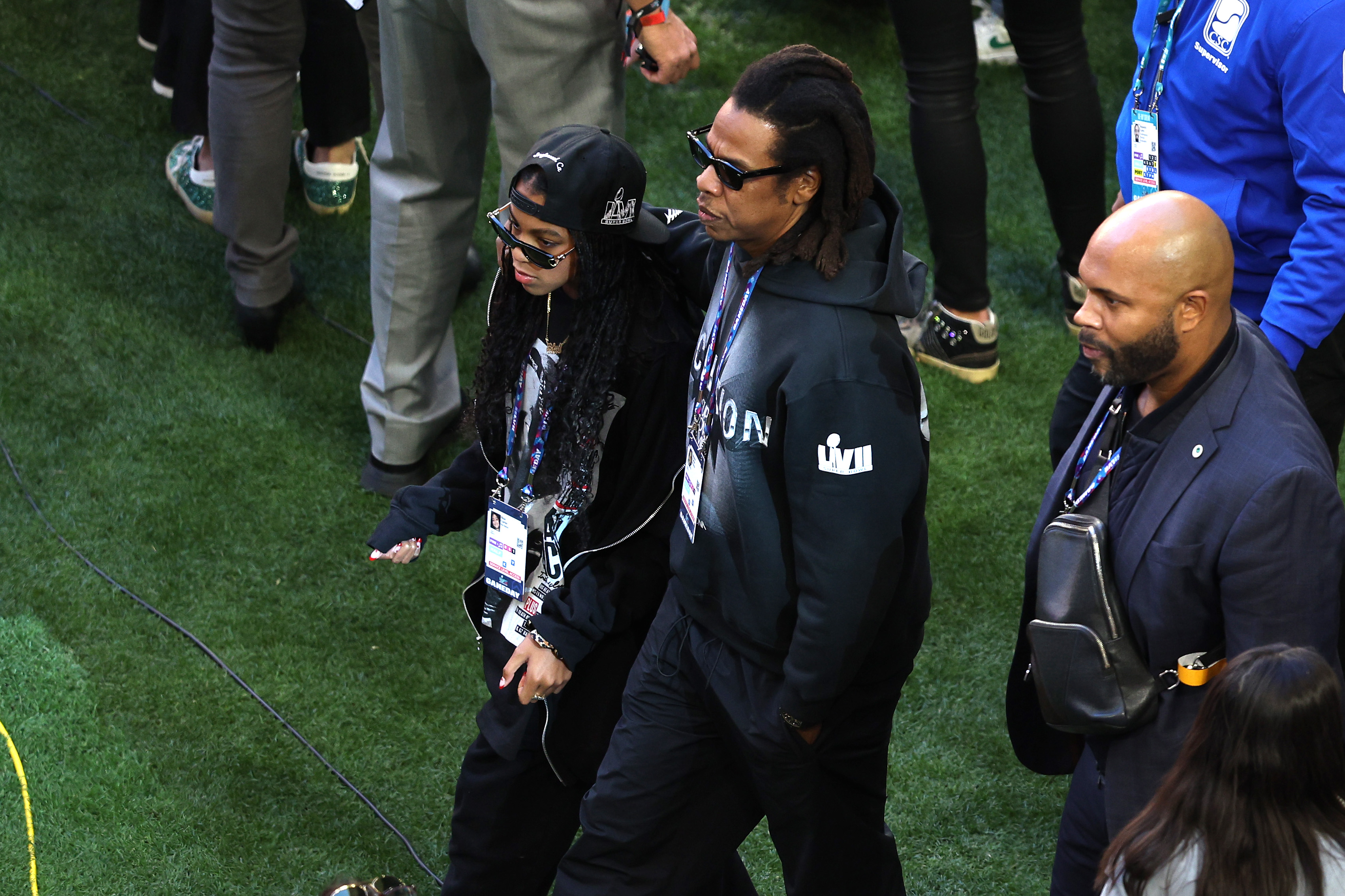 Jay-Z and Blue Ivy walk shoulder to shoulder across the field, both dressed in dark, casual layers. The image shows them moving through the crowd as pregame preparations continue.