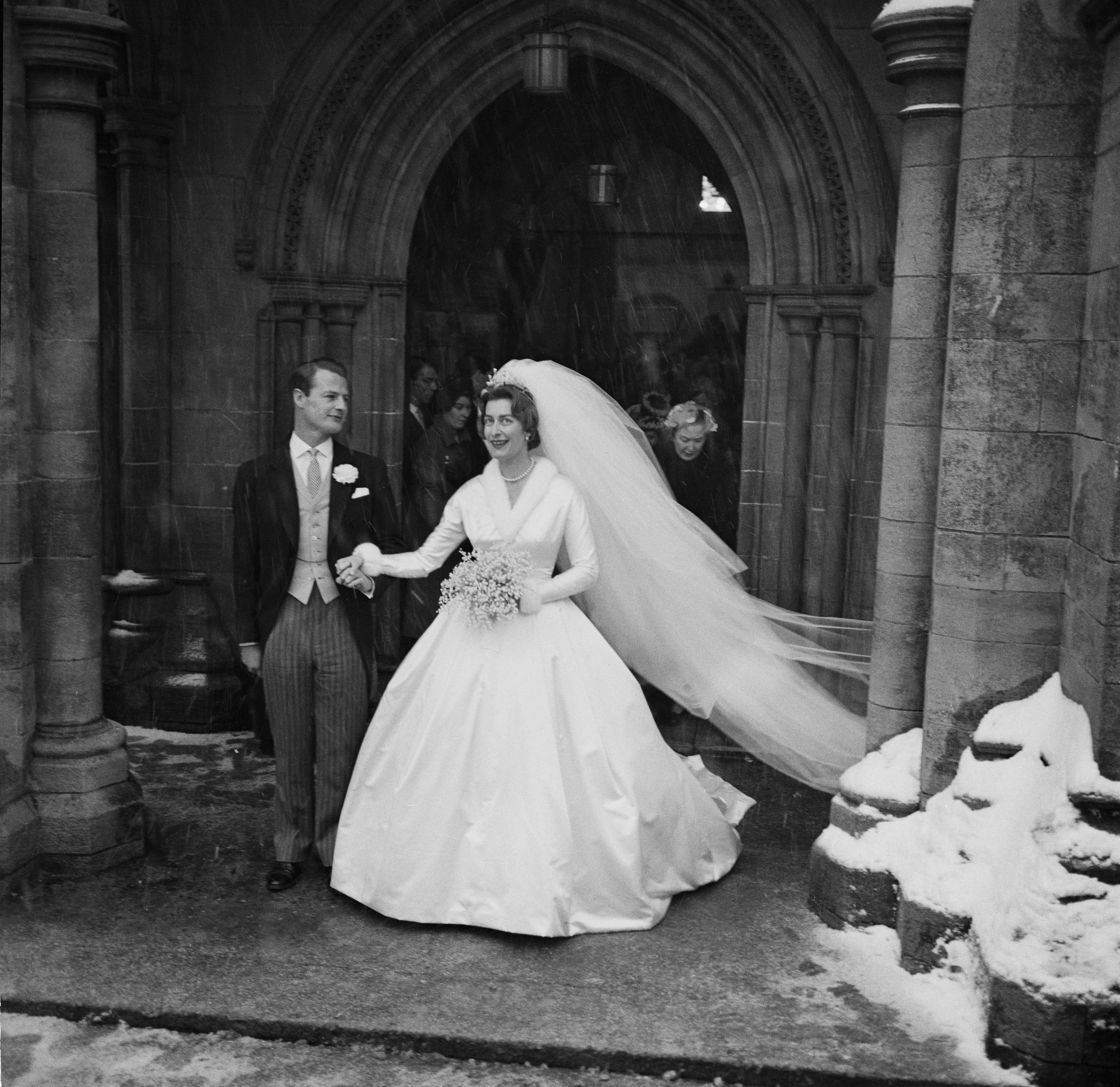Captured on 13 January 1960 at Romsey Abbey in Hampshire, Lady Pamela Mountbatten emerges in a full-skirted wedding gown and flowing veil beside David Hicks, the newlyweds stepping out into falling snow in a moment of timeless celebration outside the historic church.