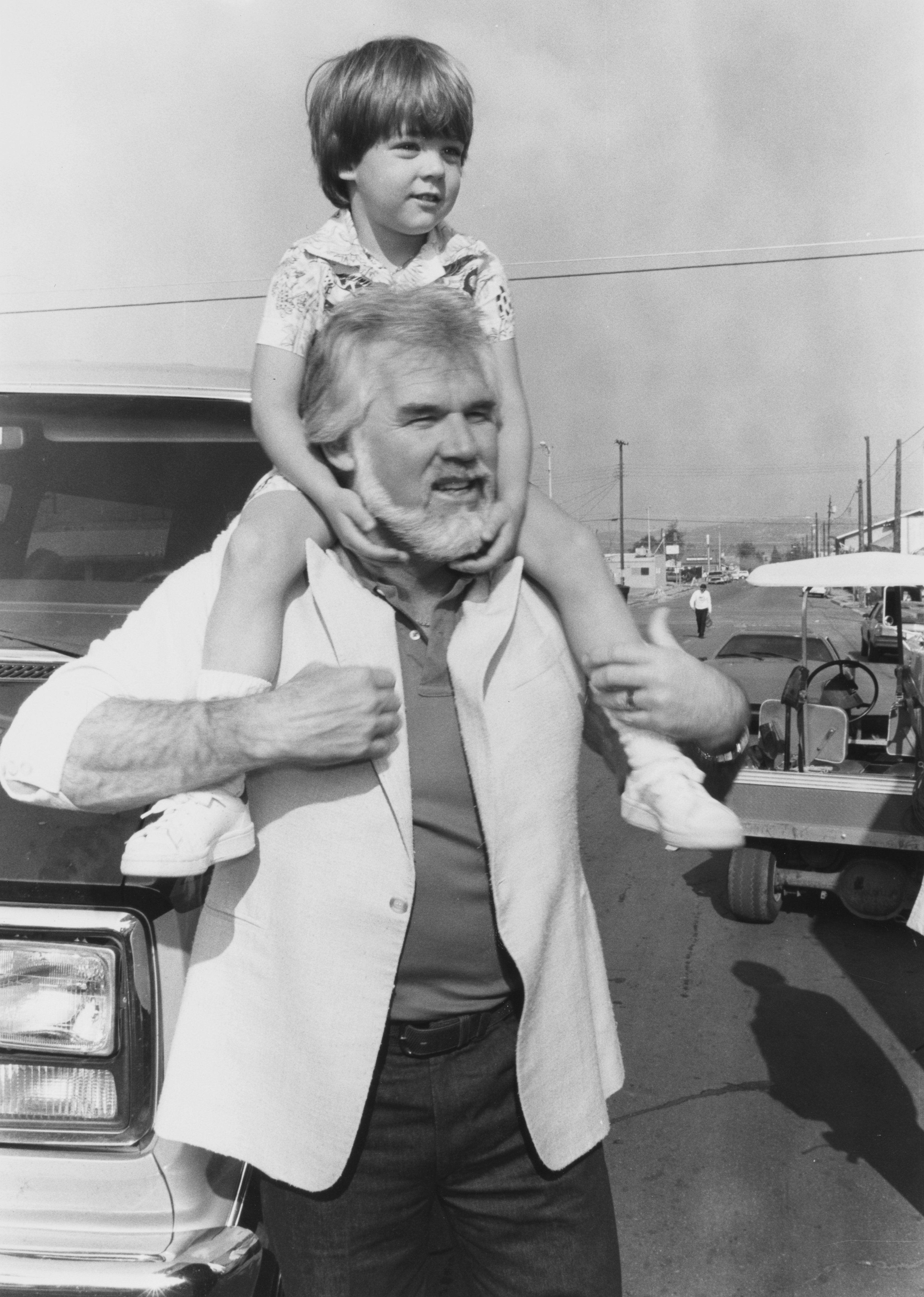 American singer-songwriter and musician Kenny Rogers (1938-2020) and his son, Christopher Rogers, who sits on his father's shoulders, attend the taping of the video and commerical for the 'Hands Across America' campaign benefit, at Center Street in Taft, California, 18th January 1986. | Source: Getty Images