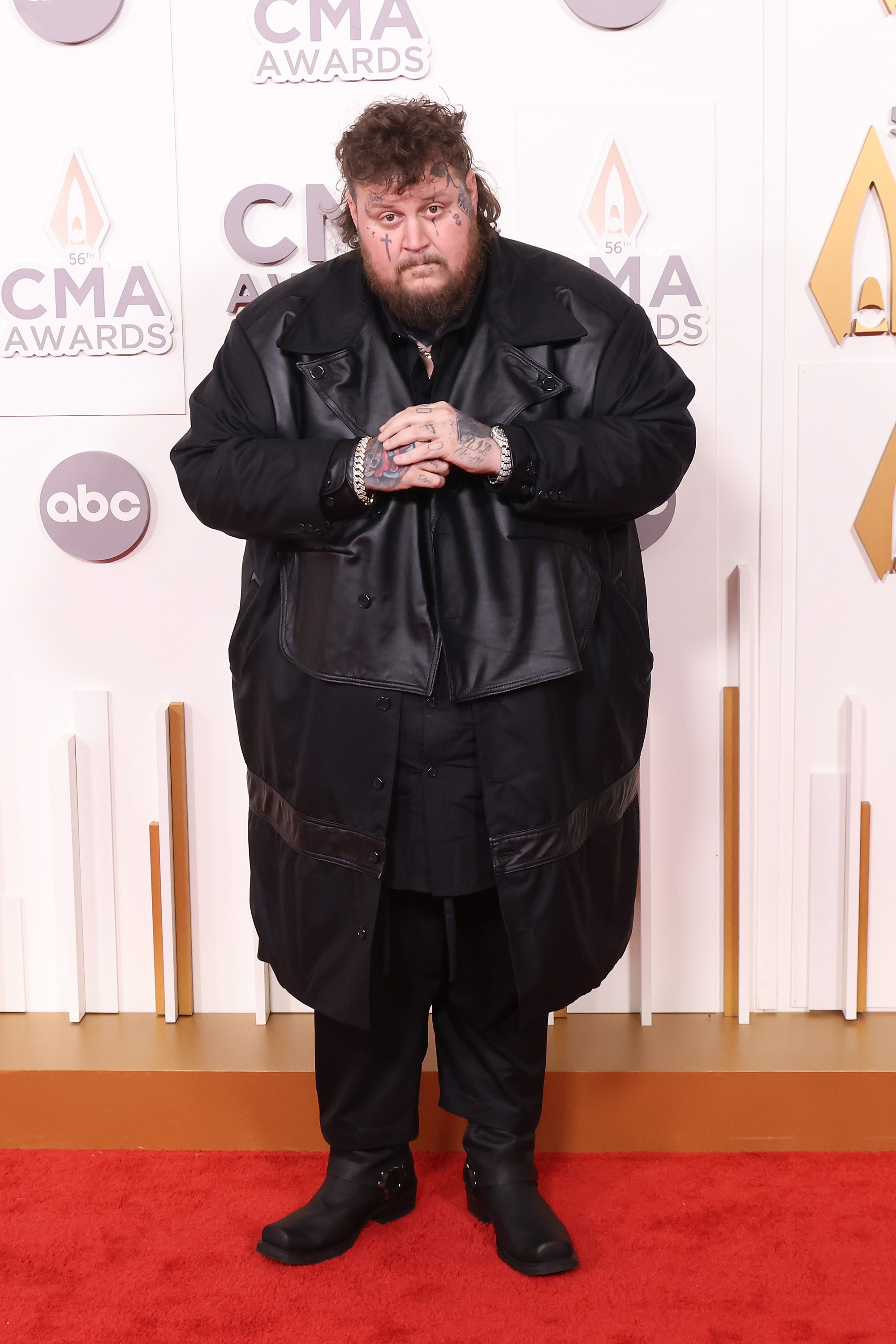 Jason "Jelly Roll" stands on the red carpet at the CMA Awards, wearing an all-black ensemble topped with a long leather coat. His look is completed with black boots, layered silver chains, and his signature face tattoos, giving him a bold and distinctive presence. With his hands clasped together, he strikes a serious yet confident pose for the cameras.