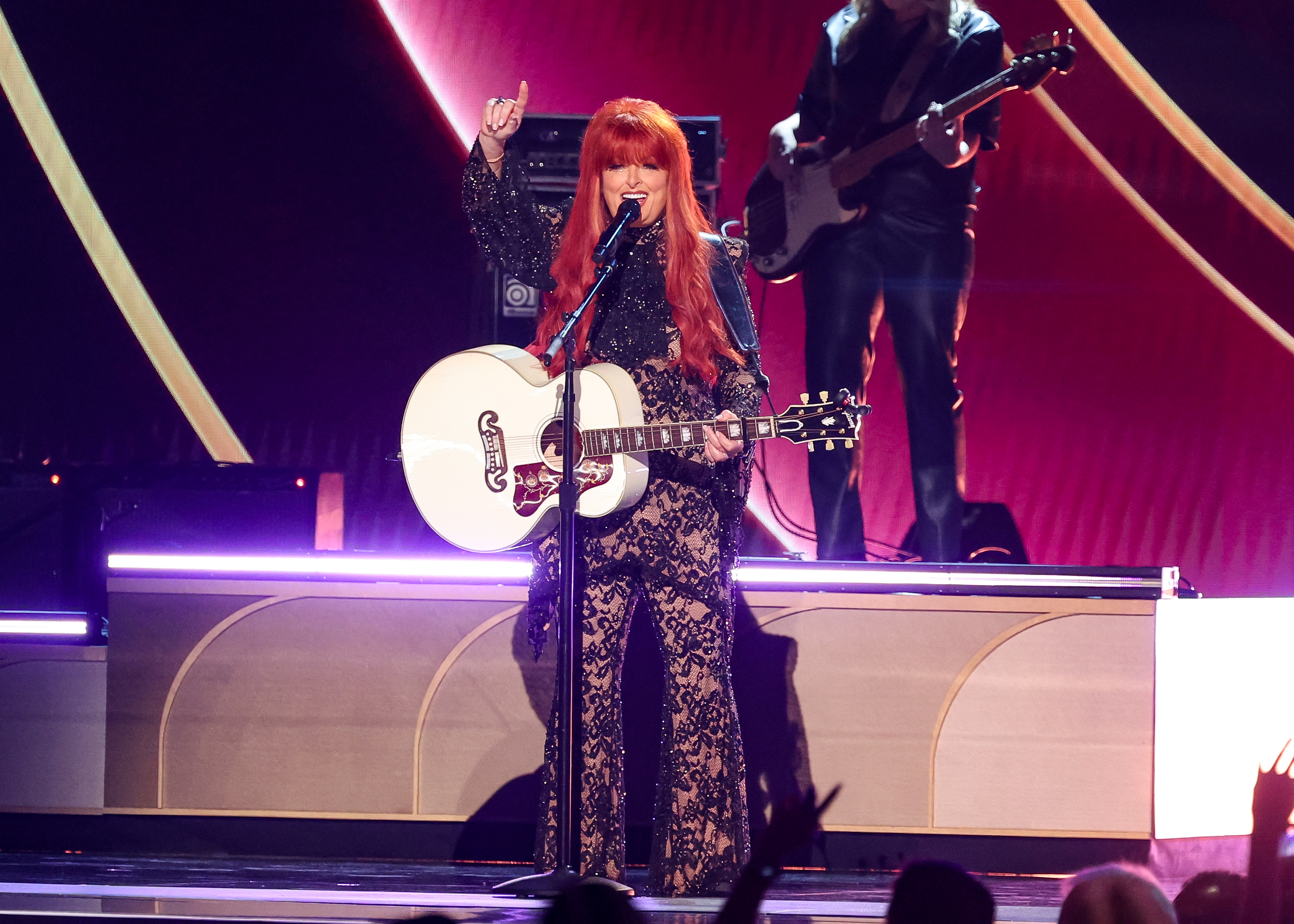 Wynonna Judd performs onstage at the 60th ACM Awards in Frisco, Texas, wearing her glittering lace look while playing a white guitar. Her high-energy performance was a highlight of the evening, drawing cheers from the crowd.