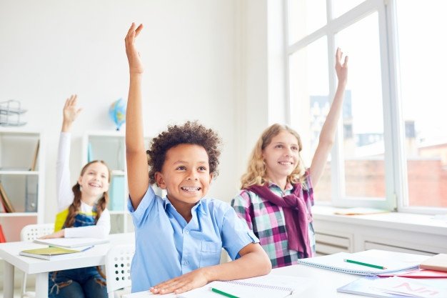 Photo of young children raising their hands in a classroom. | Photo: Freepik