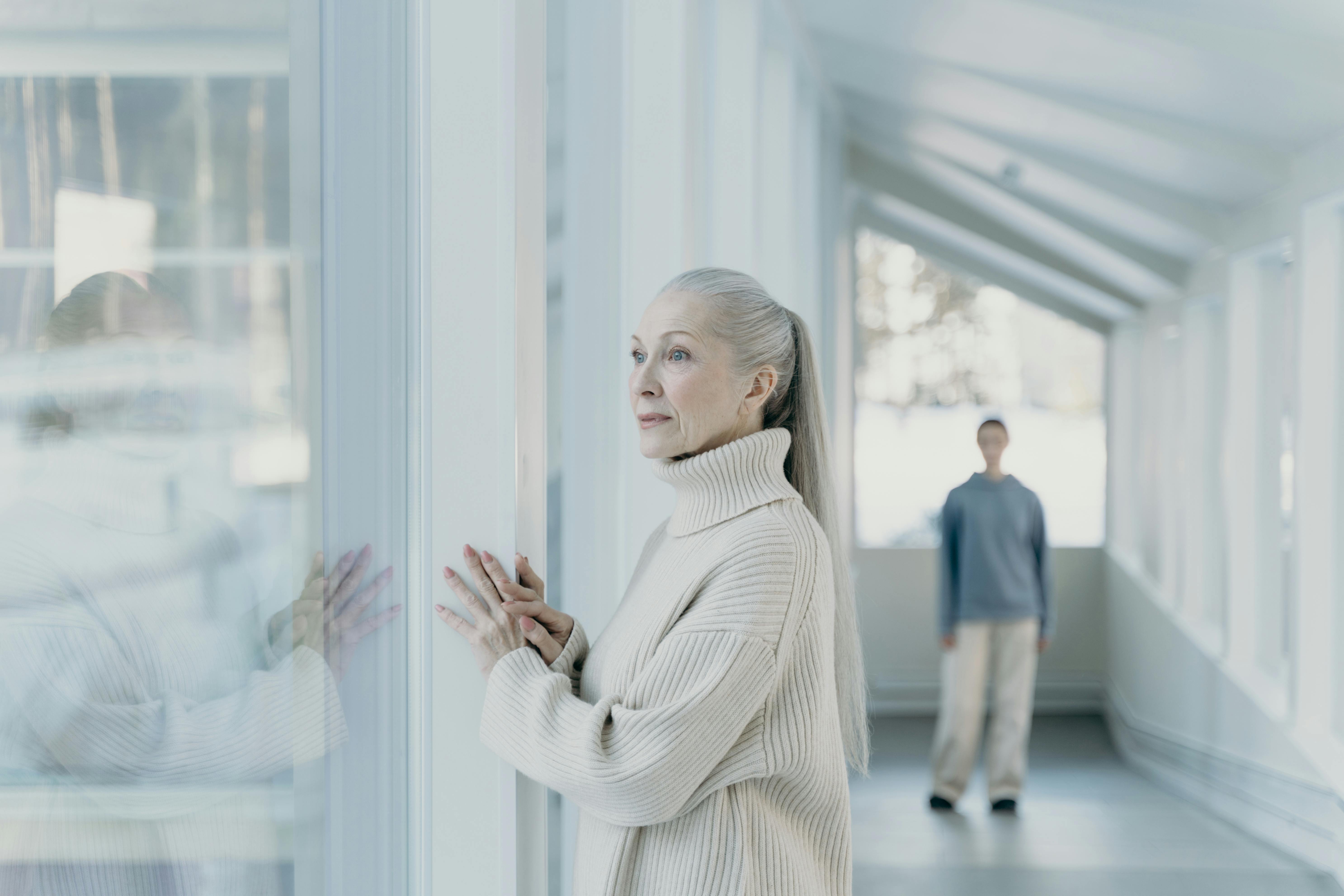 A elderly woman looks through window glass, with a figure in the background | Source: Pexels
