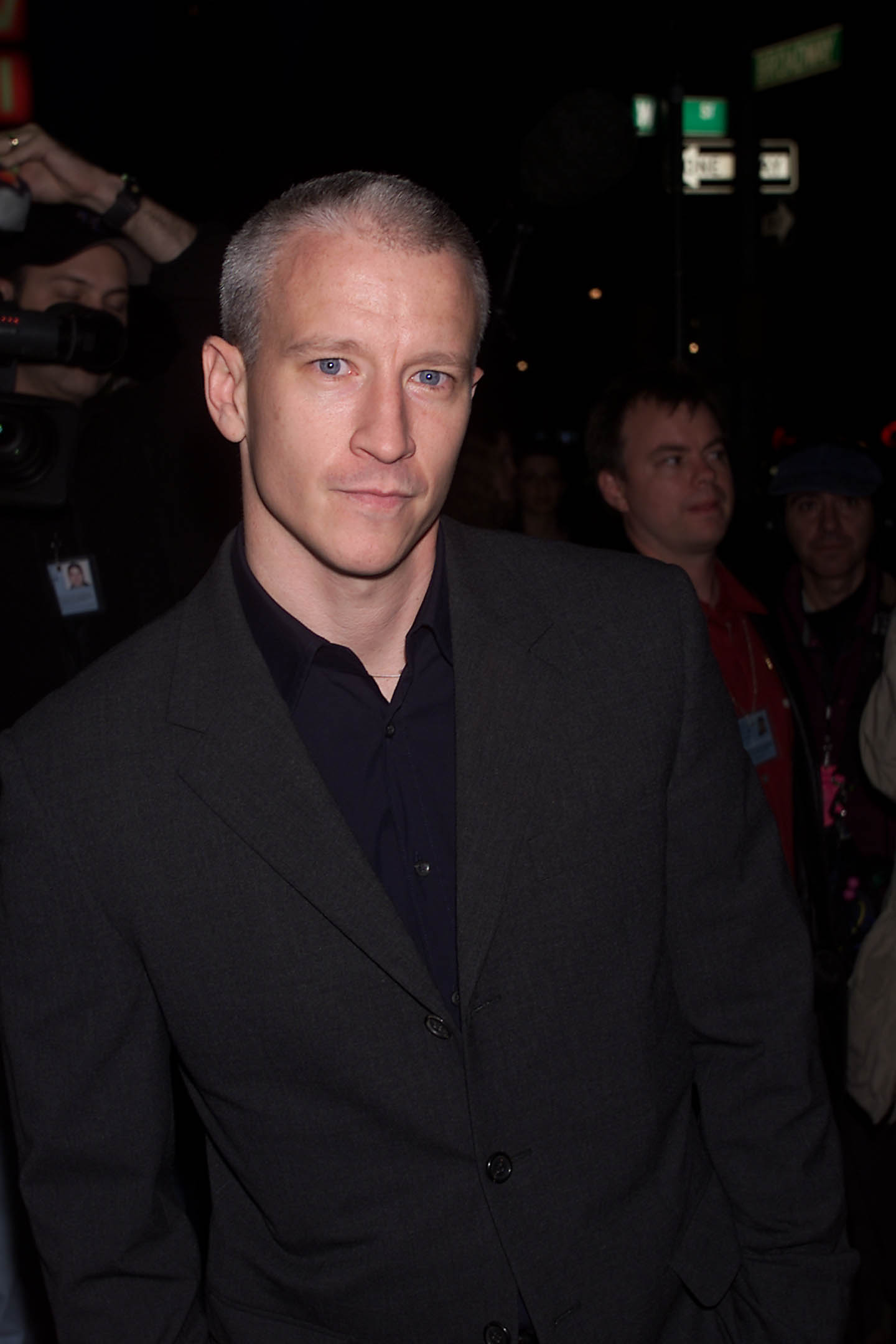 A young Anderson Cooper attends a nighttime event in New York City, sporting a close-cropped haircut and dressed in a charcoal blazer over a dark shirt, his blue eyes standing out under the city lights.