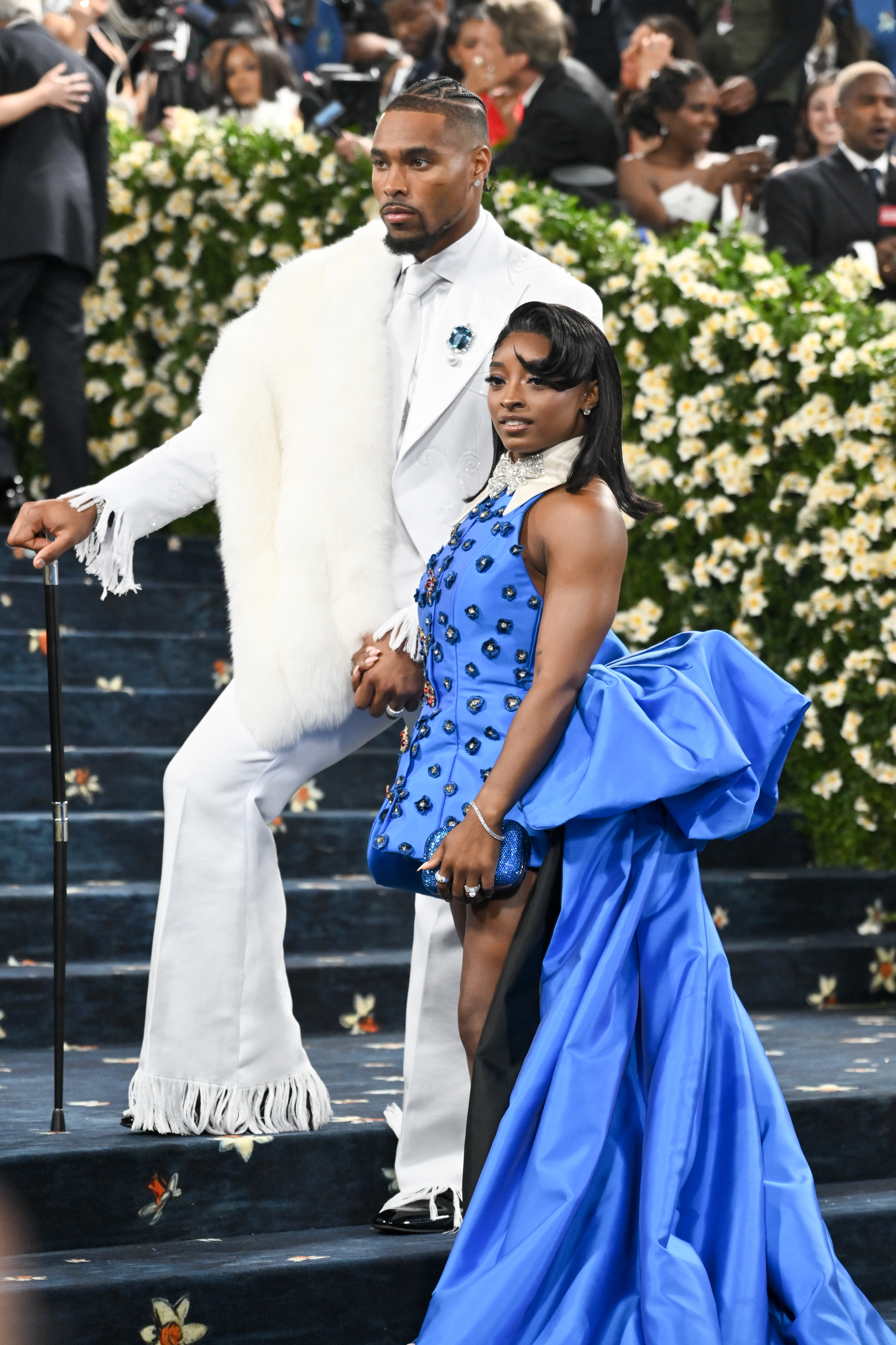 Jonathan Owens holds Simone Biles' hand as they ascend the steps. Both wear composed expressions as they navigate the crowd under flashing lights.