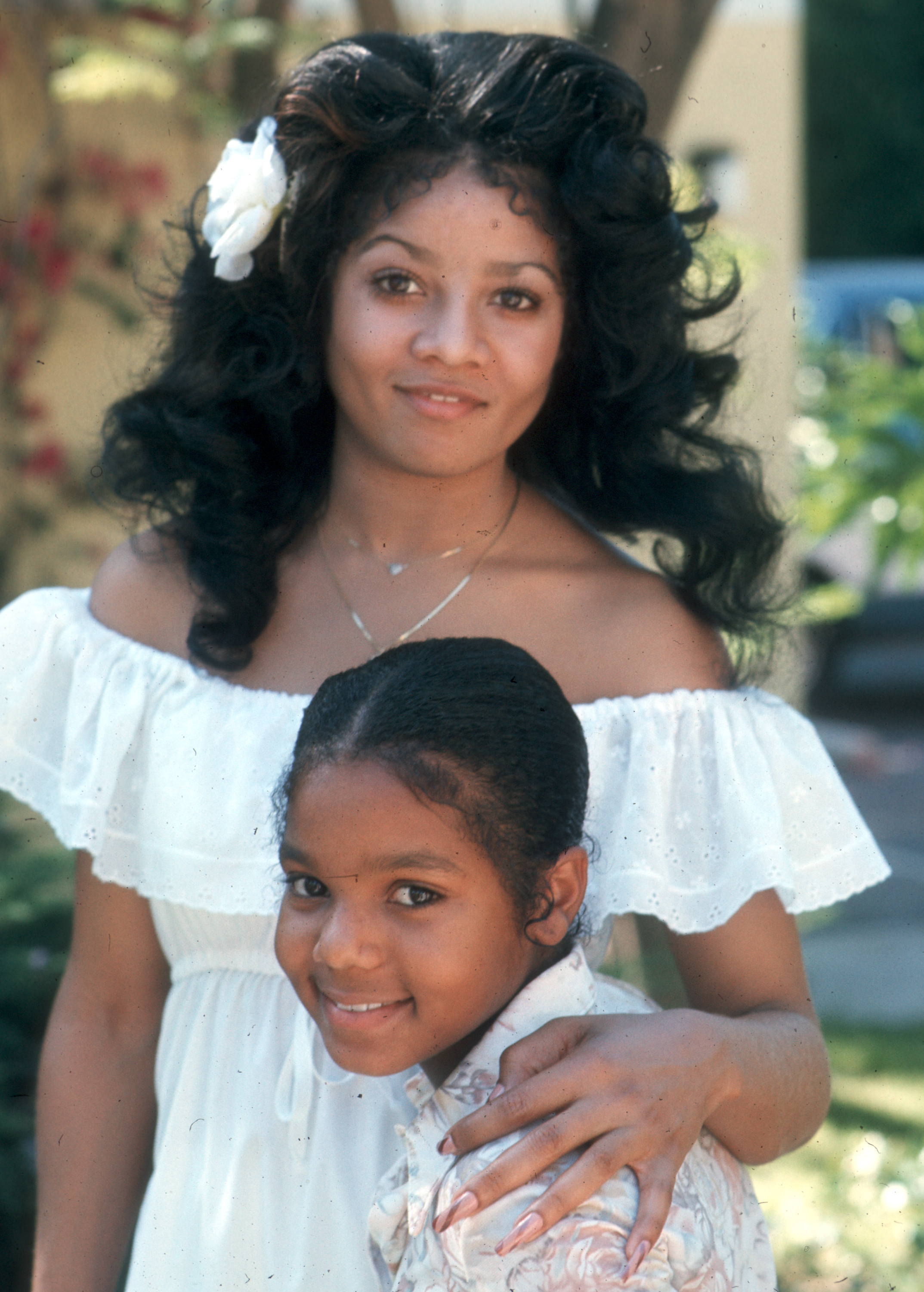 In a sunlit portrait session, sisters La Toya and Janet Jackson share a sweet moment in Los Angeles. La Toya, wearing an off-the-shoulder white dress and a flower in her hair, exudes soft elegance as she wraps her arm around a young Janet.