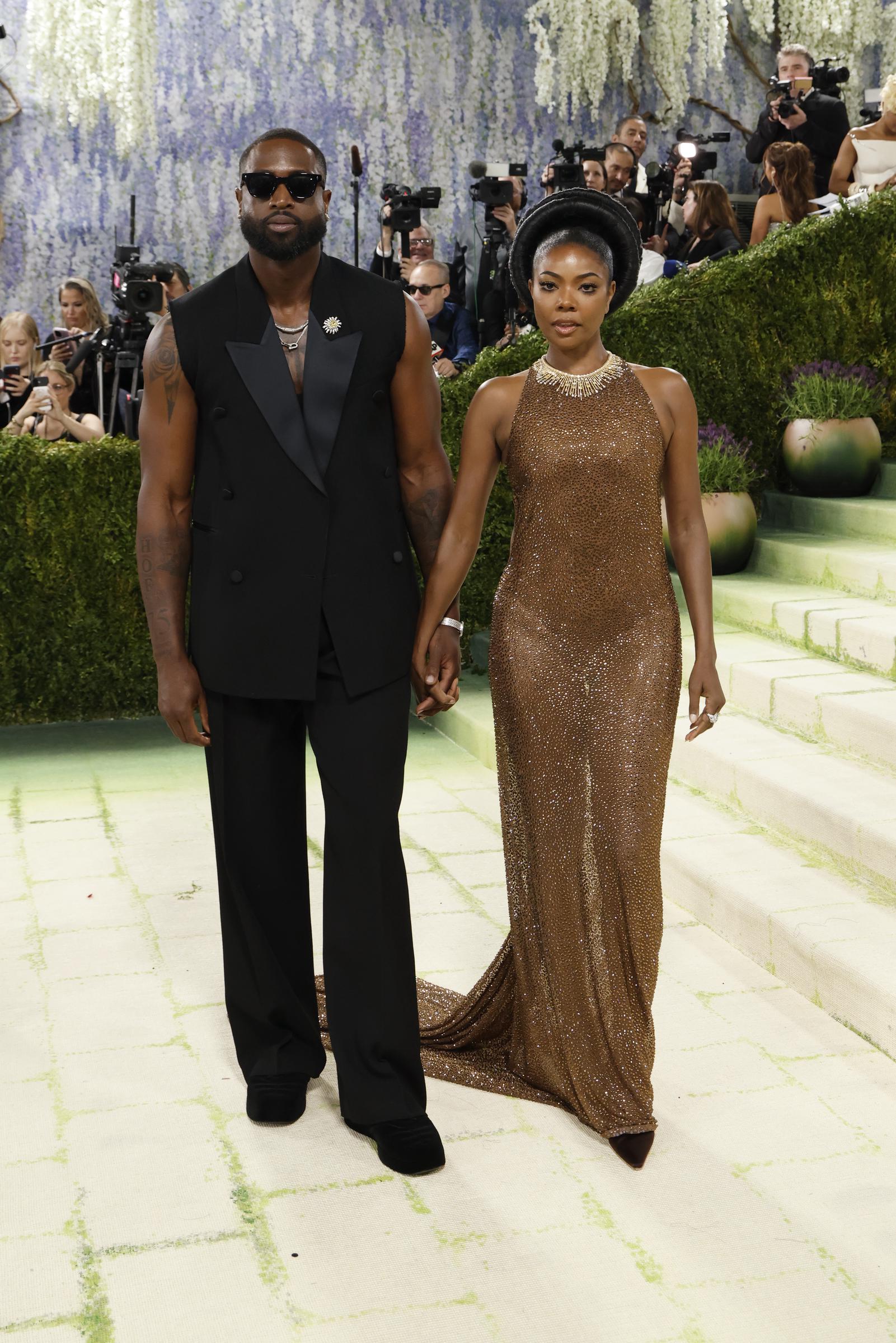 Dwyane Wade stands in a tailored sleeveless black look with dark sunglasses and layered jewelry. Beside him, Gabrielle Union shines in a form-fitting bronze gown, the two holding hands in a coordinated, confident pose.