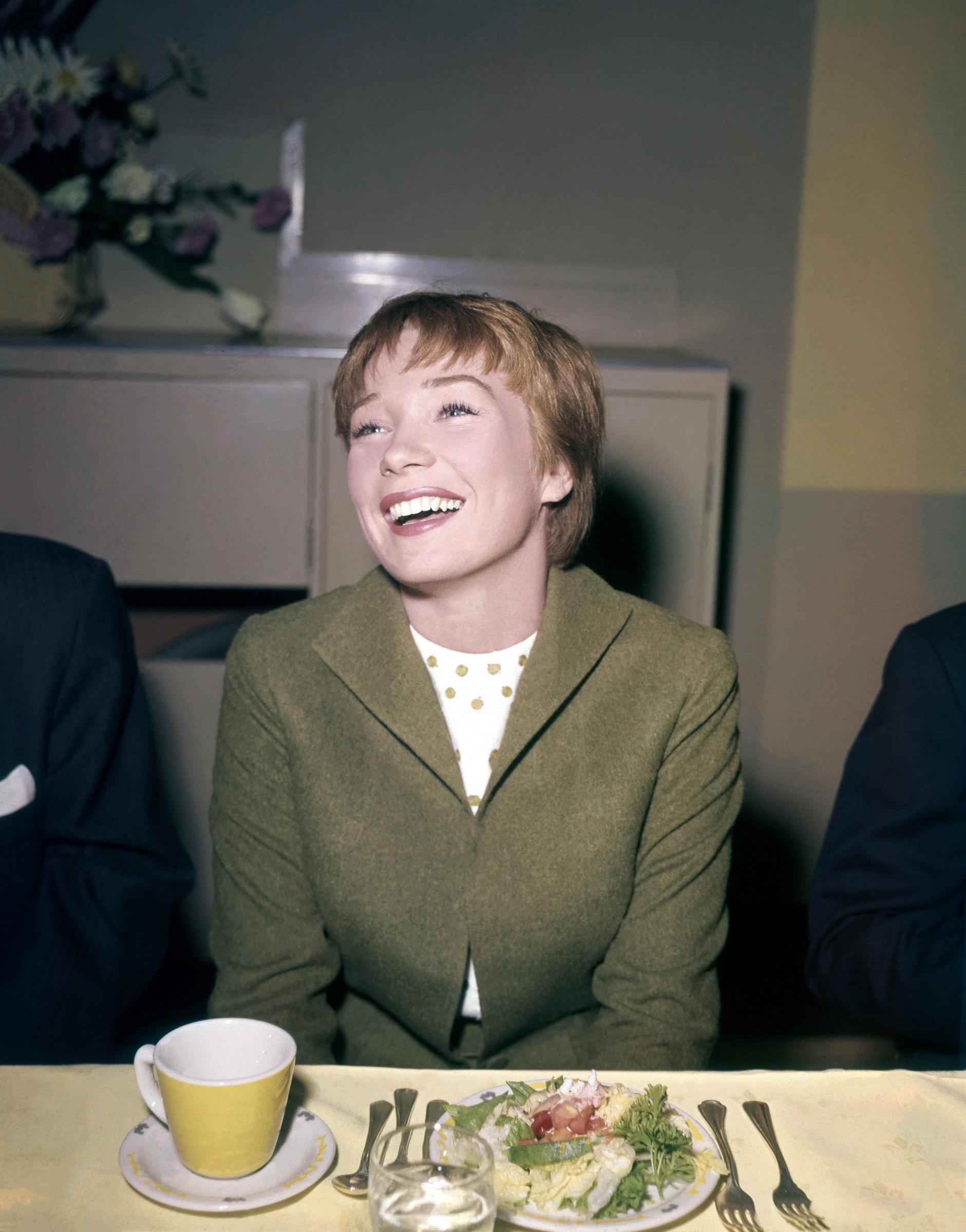 A young Shirley MacLaine lights up the room during a lunch in Los Angeles, circa 1959. Draped in a smart green jacket and patterned blouse, the rising star is caught mid-laughter — her charm and charisma already undeniable as she soared to Hollywood stardom.