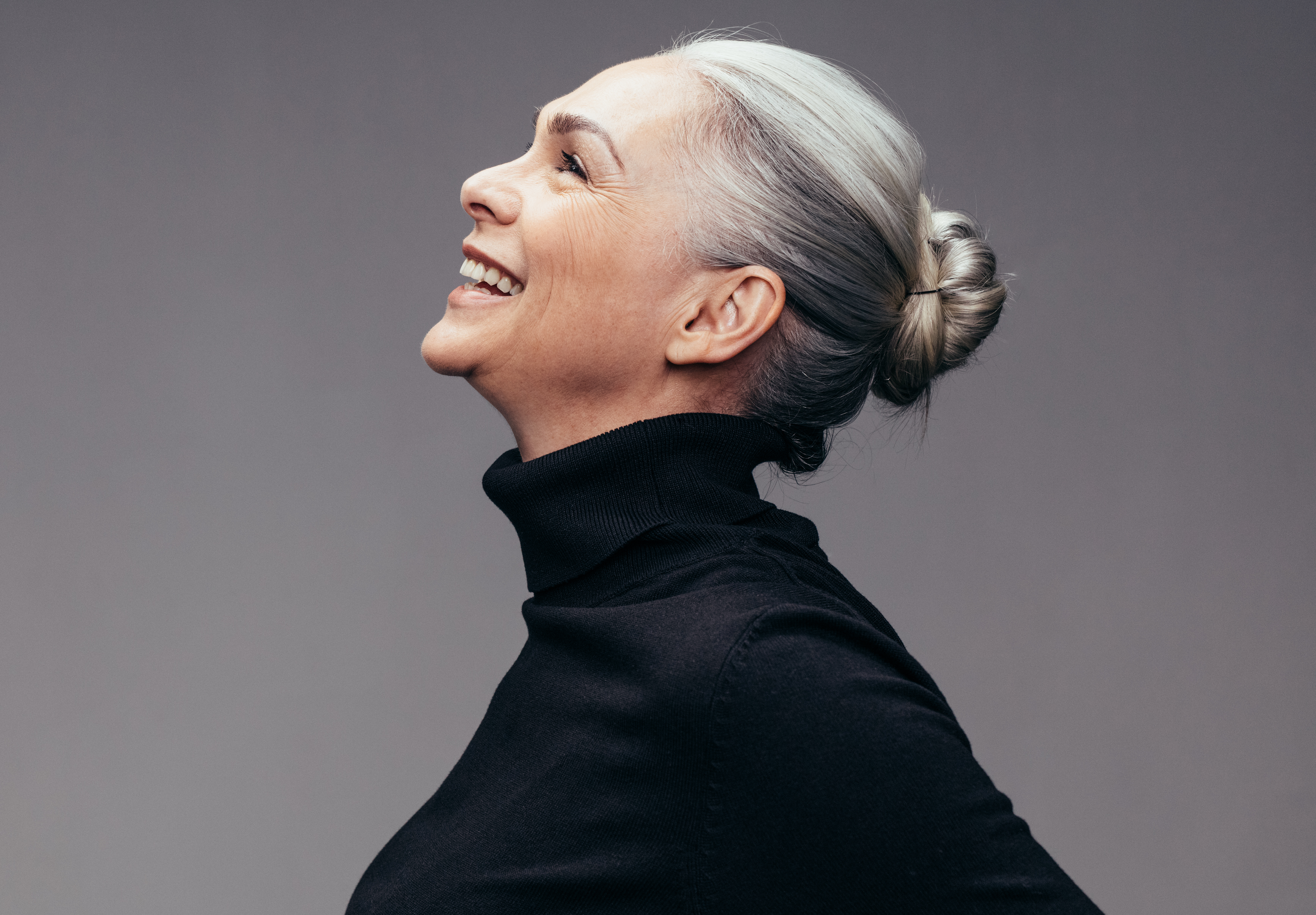 Grey-haired woman with her hair in a bun tilting her head back and laughing | Source: Shutterstock