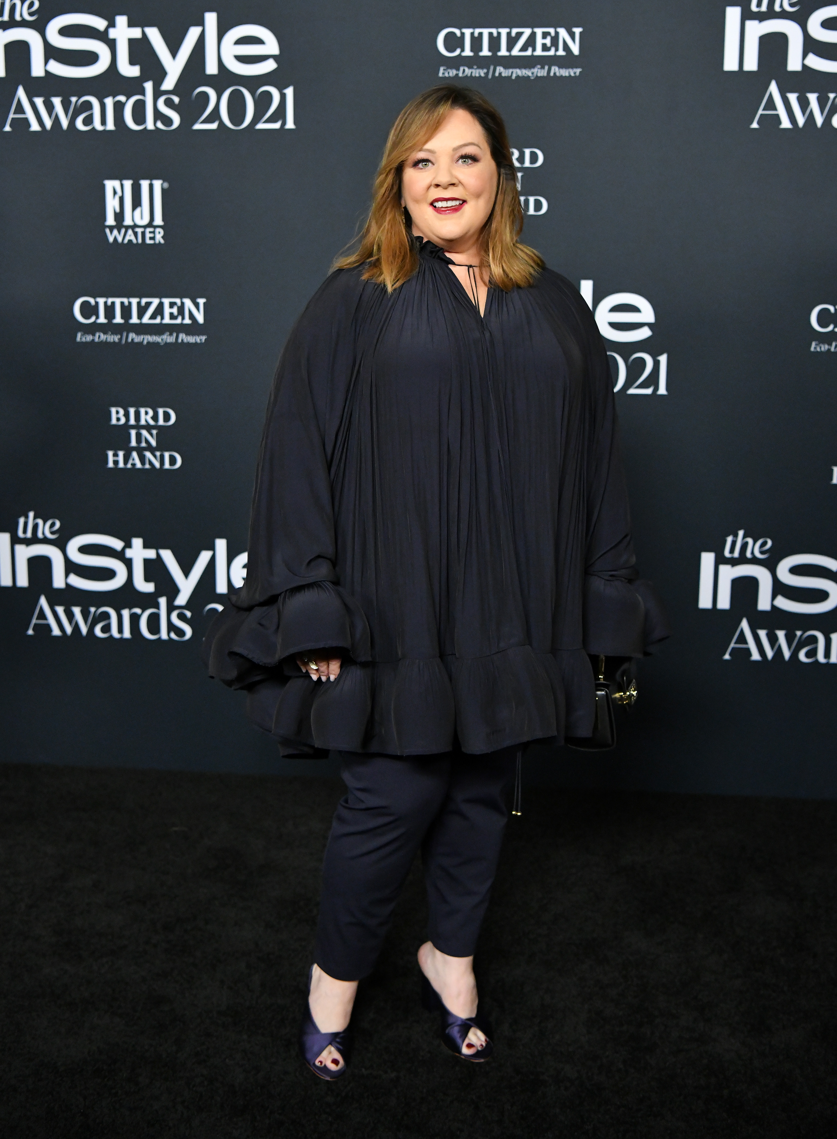 Melisa McCarthy appears poised on the black carpet, wearing a voluminous midnight-toned ensemble that adds drama and elegance to her look. With a bright smile and polished styling, she brings confidence and charm to the InStyle Awards.