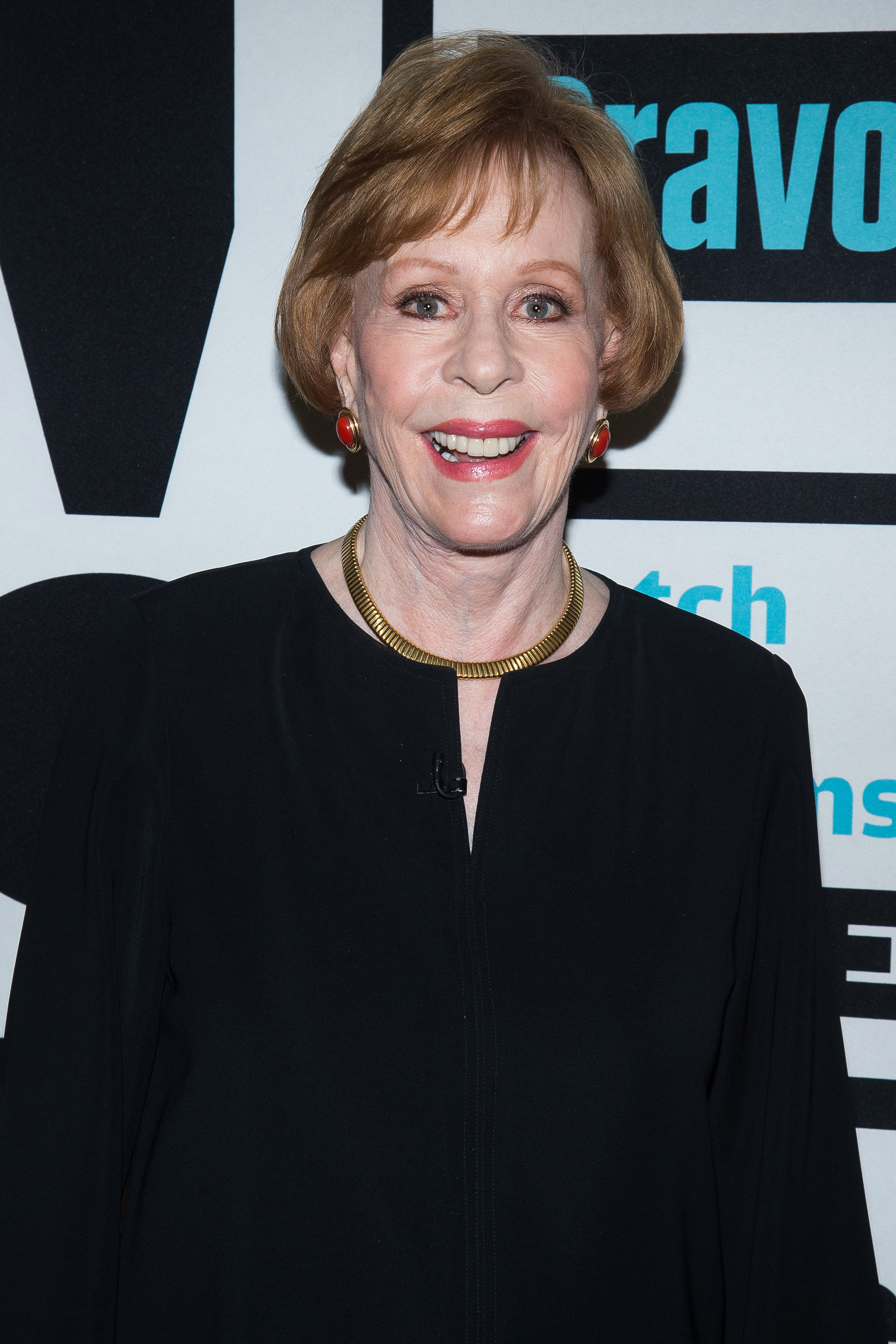 In a close-up event setting, she wears a black outfit with a gold necklace and matching red-and-gold earrings. Her hair is neatly styled, and she faces forward with a slight smile, standing against a backdrop with printed logos.
