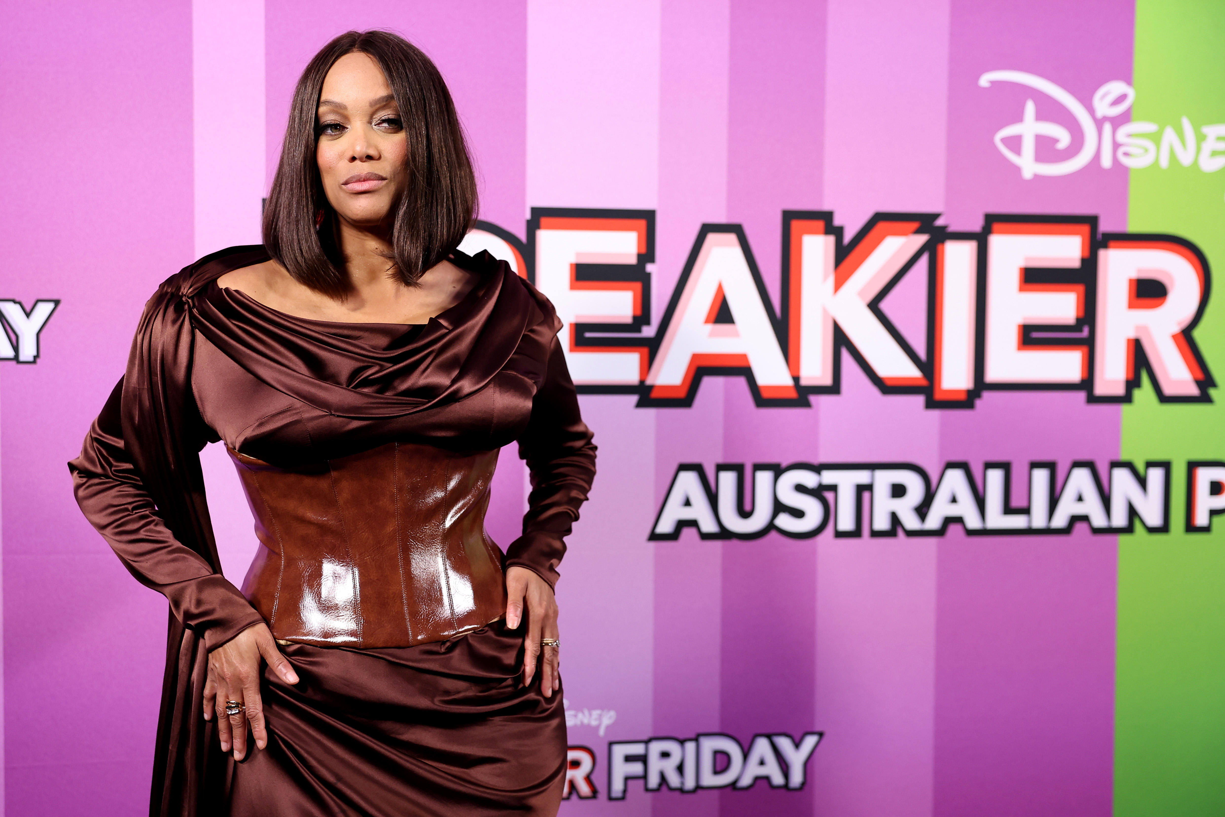 On the purple carpet, Tyra Banks strikes a confident pose, hands on hips, locking eyes with the cameras in a display of poise and self-assurance.