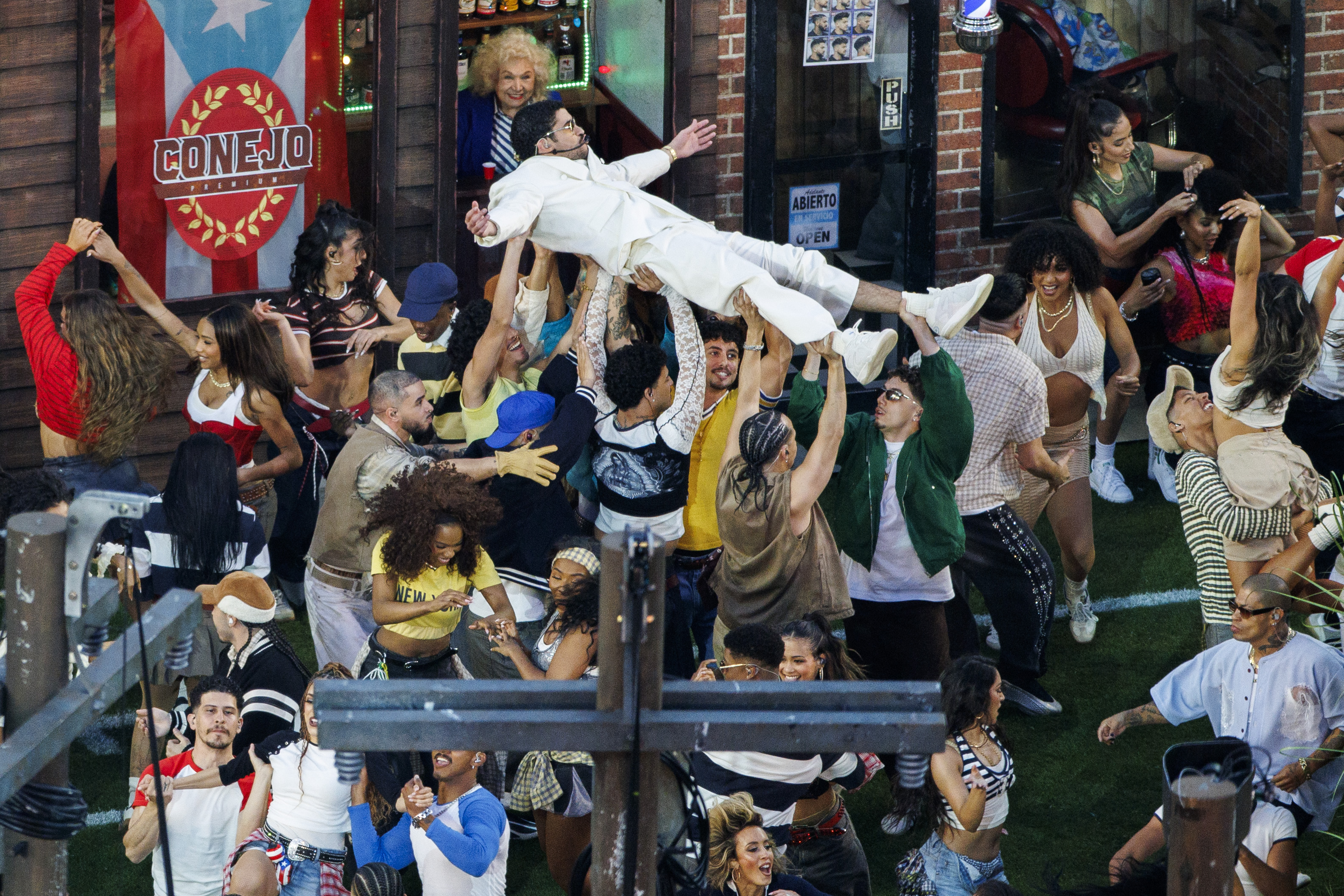 Bad Bunny is lifted by dancers while performing during the Super Bowl LX halftime show, surrounded by a crowd of performers in coordinated staging and movement.