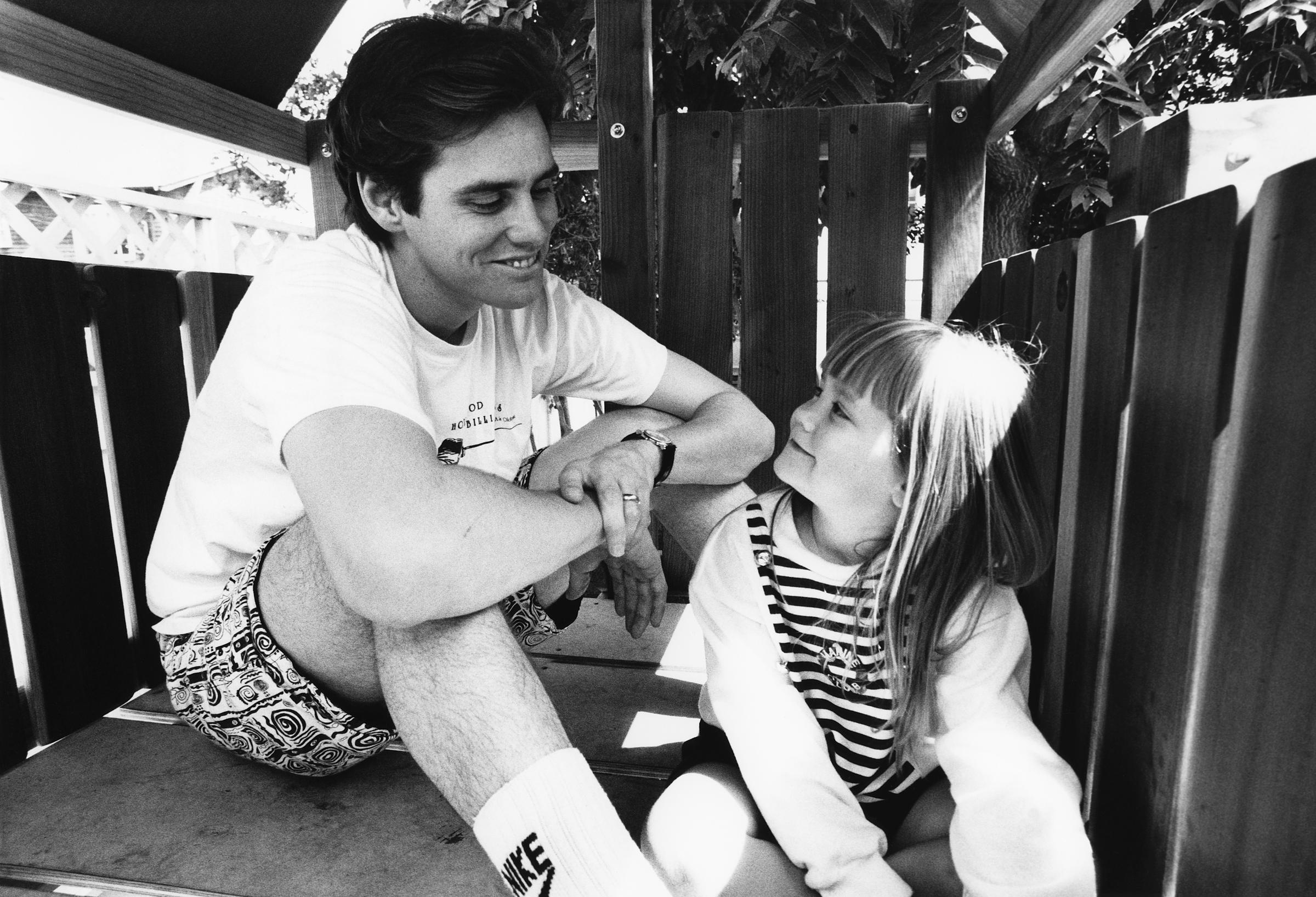 The black-and-white photo captures Jim Carrey sharing a tender, joyful moment with his daughter as they sit together in a playhouse. Both are smiling warmly, their eyes meeting in a look of pure affection and connection. The relaxed, candid setting highlights their loving father-daughter bond and genuine happiness.