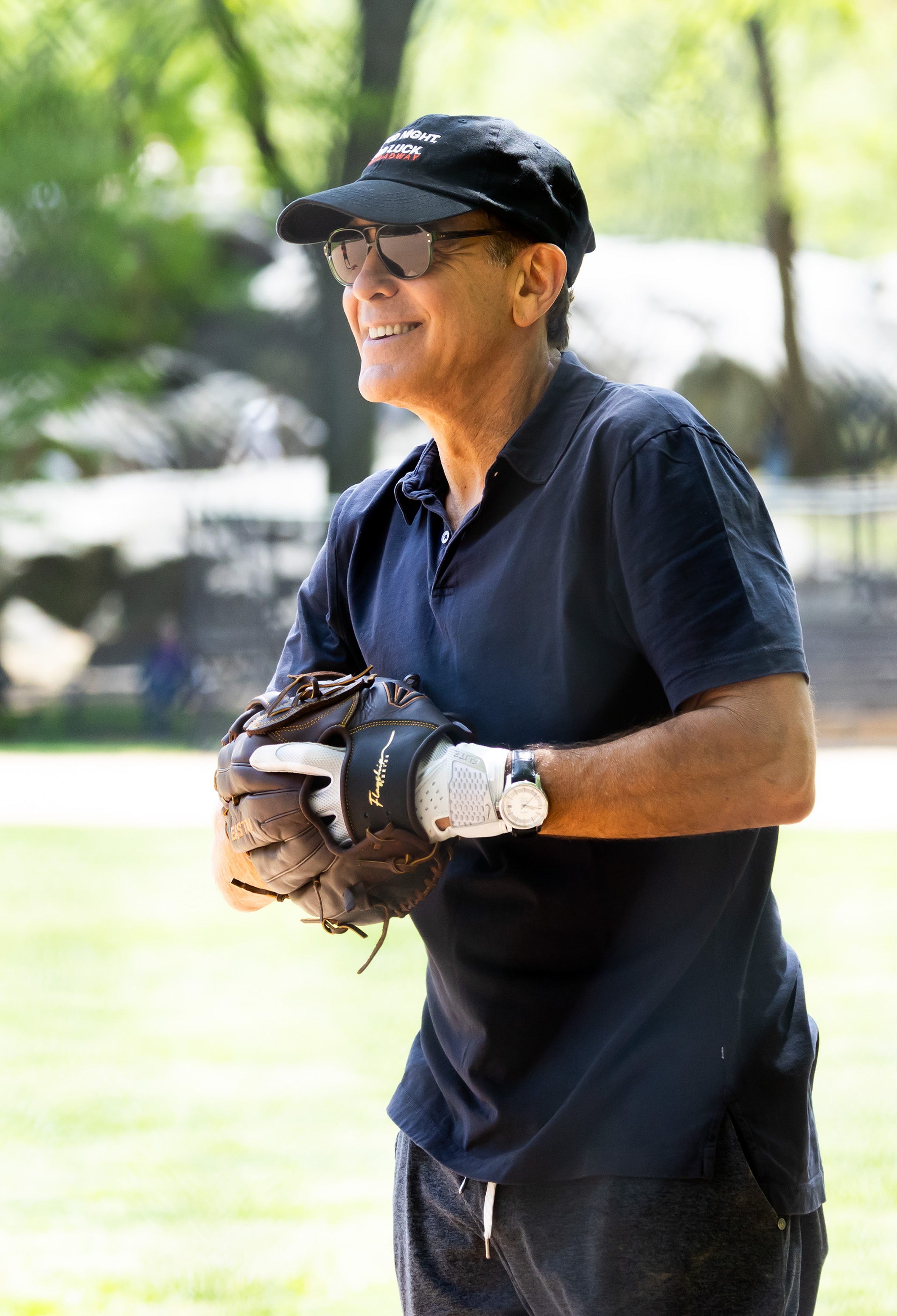 Clooney smiles mid-game, dressed in a dark polo with a baseball glove in hand. Sporting sunglasses and a black cap that reads “Good Night, and Good Luck Broadway,” he looks relaxed and focused, bringing his trademark charm to the ball field.